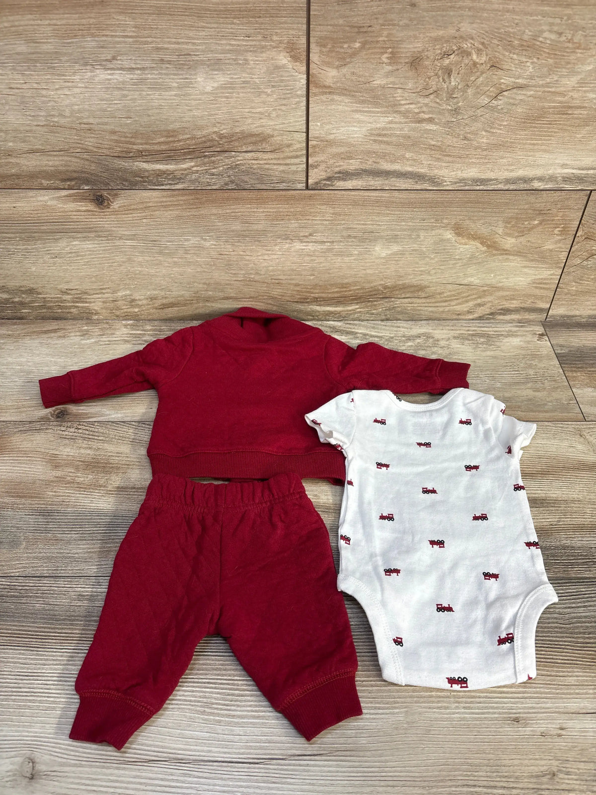 Top view of Carter’s three-piece newborn set with red quilted jacket and pants, white printed bodysuit against wood flooring