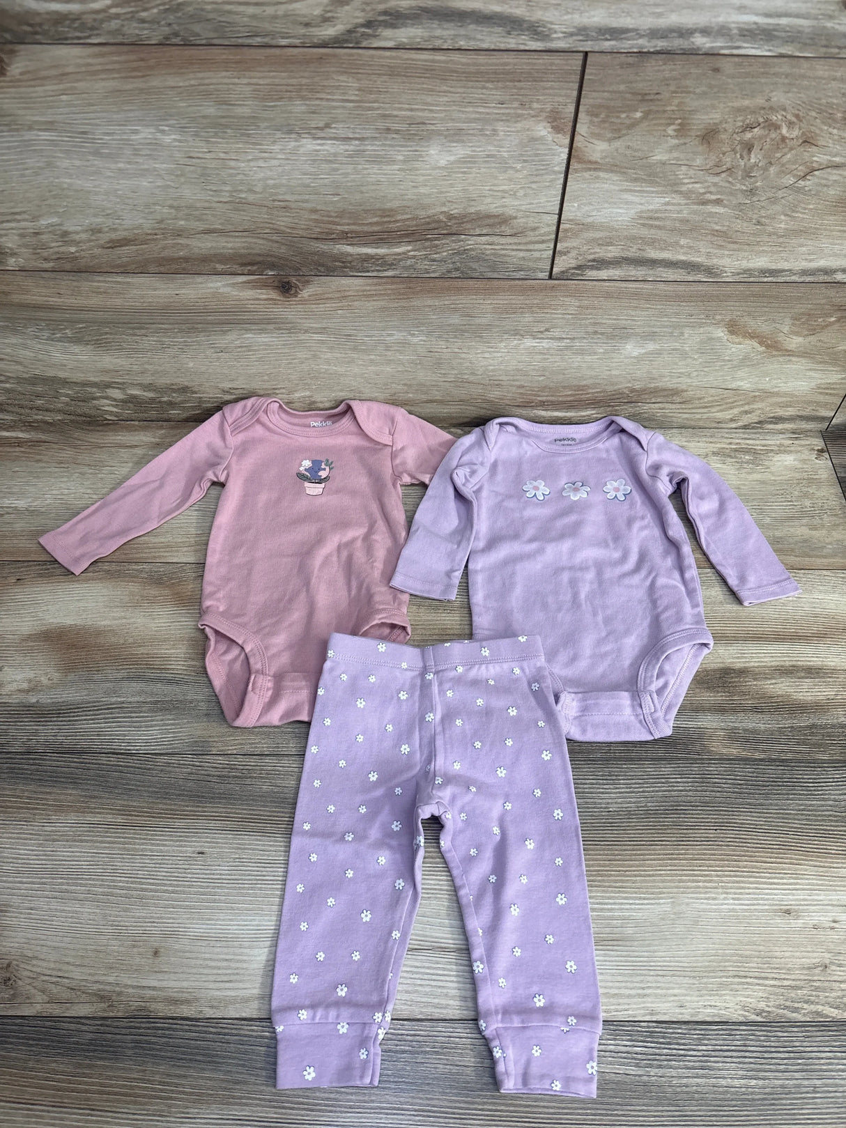 Flat lay of Pekkle 3pc Earth bodysuit set 3-6m: pink and lavender bodysuits with floral appliques and lavender daisy leggings on wood