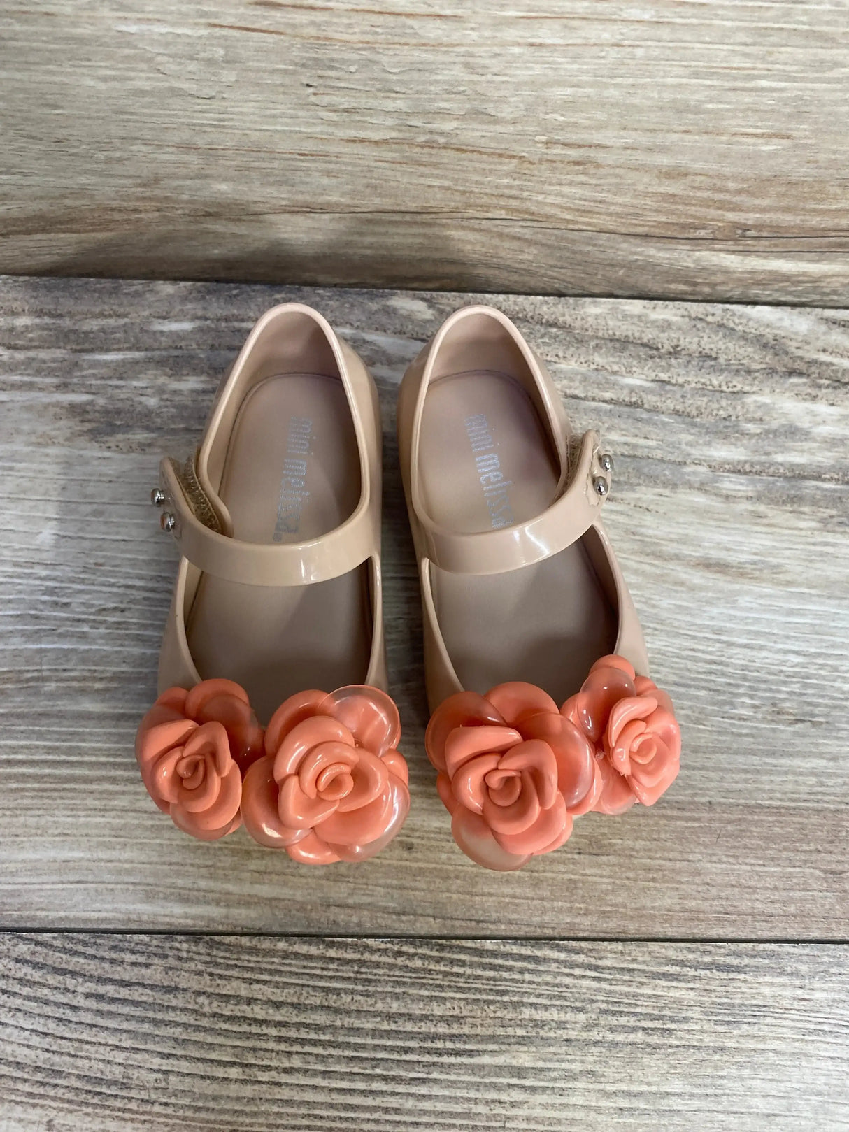Top view of Mini Melissa Ultragirl beige-pink flats showing coral flower embellishments and Mary Jane strap on wood background