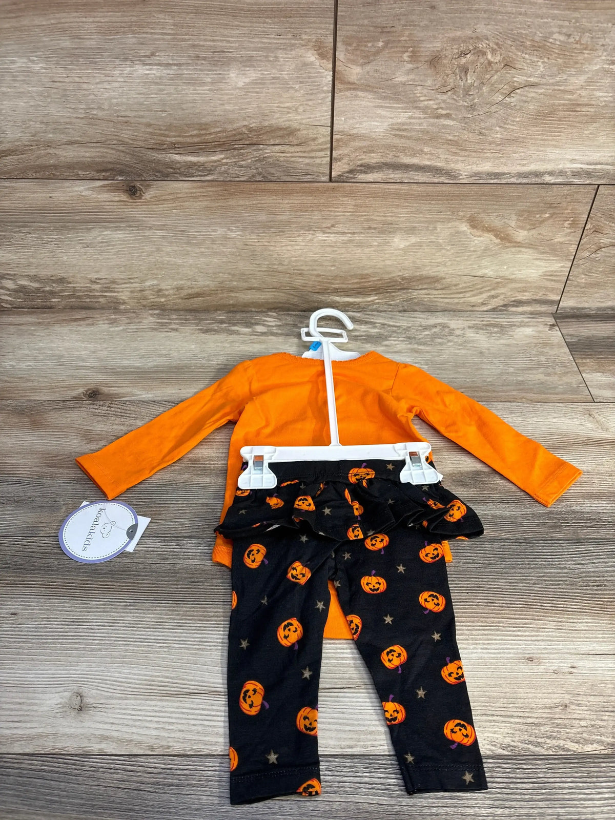 Front view of orange baby bodysuit and black pants set showcasing pumpkin and star print on a wood floor