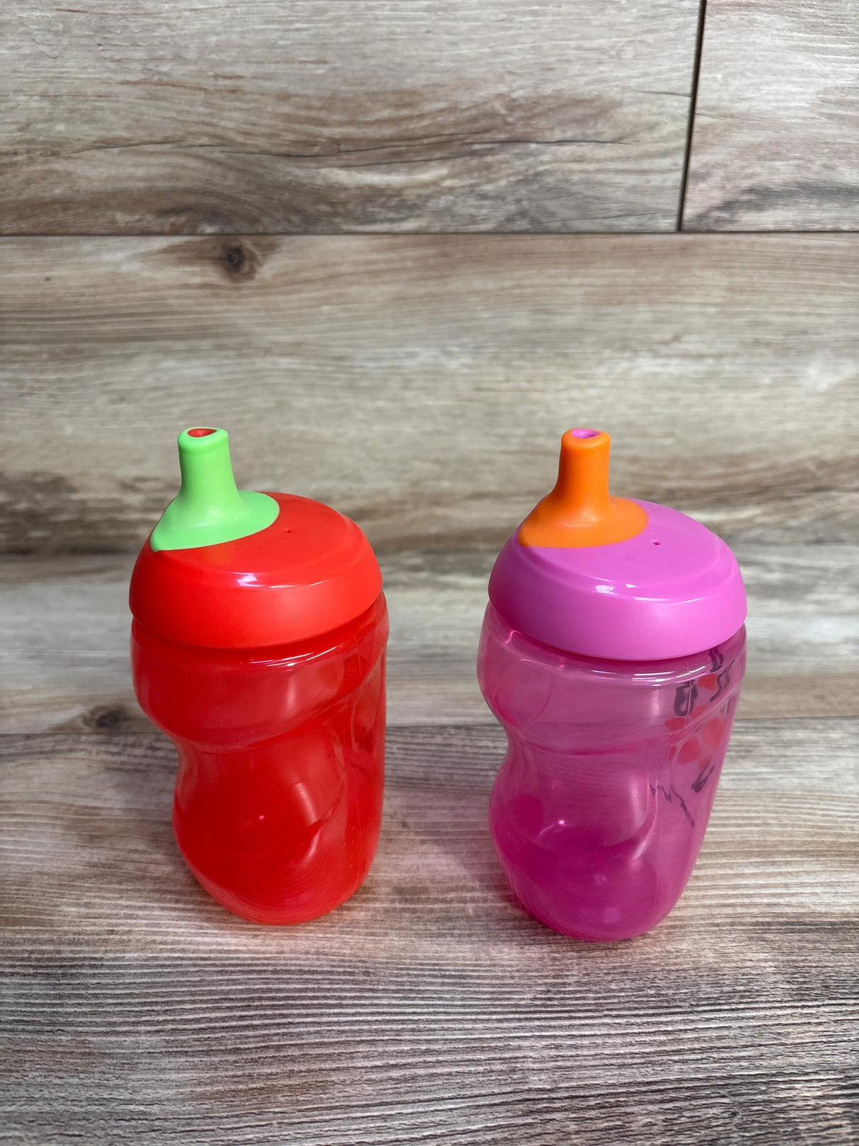 Red and pink plastic sippy cups with colorful lids on wood background