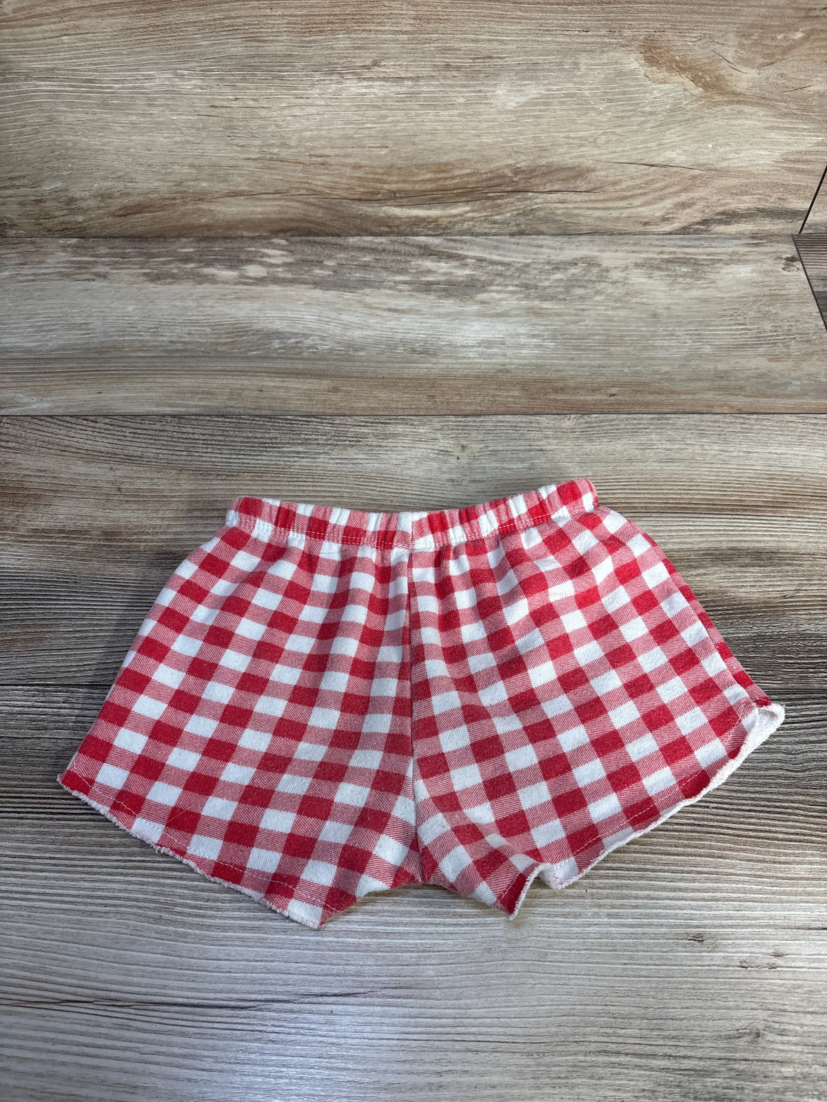 Flat-lay view of Grayson Mini checker shorts in size 3T showcasing red and white checker pattern on a wood-grain background