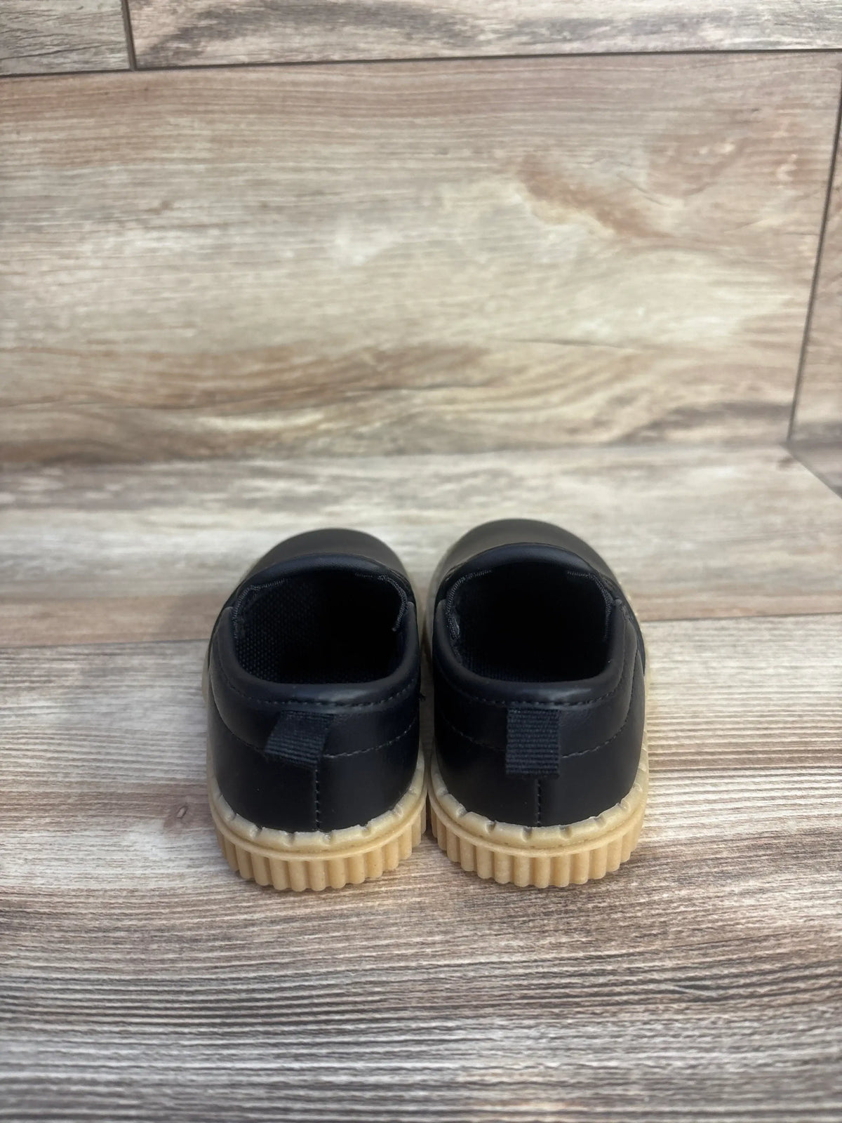 Rear view of OshKosh Victor slip-on sneakers showcasing black uppers and beige ribbed soles on a wood-look floor