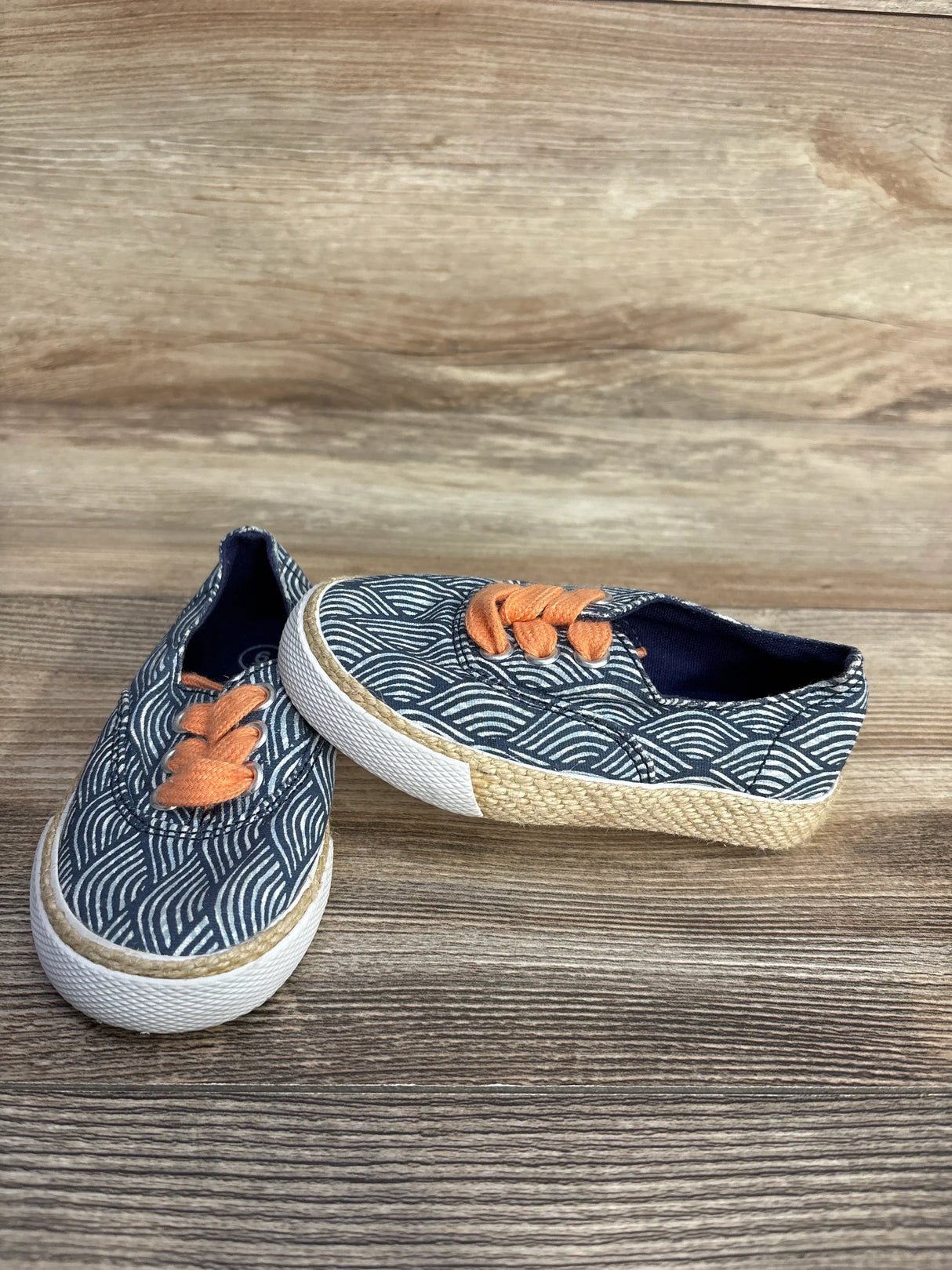 Three-quarter view of Genuine Kids OshKosh Tailynn slip-on sneakers in navy wave print with orange lobster patch on wood floor