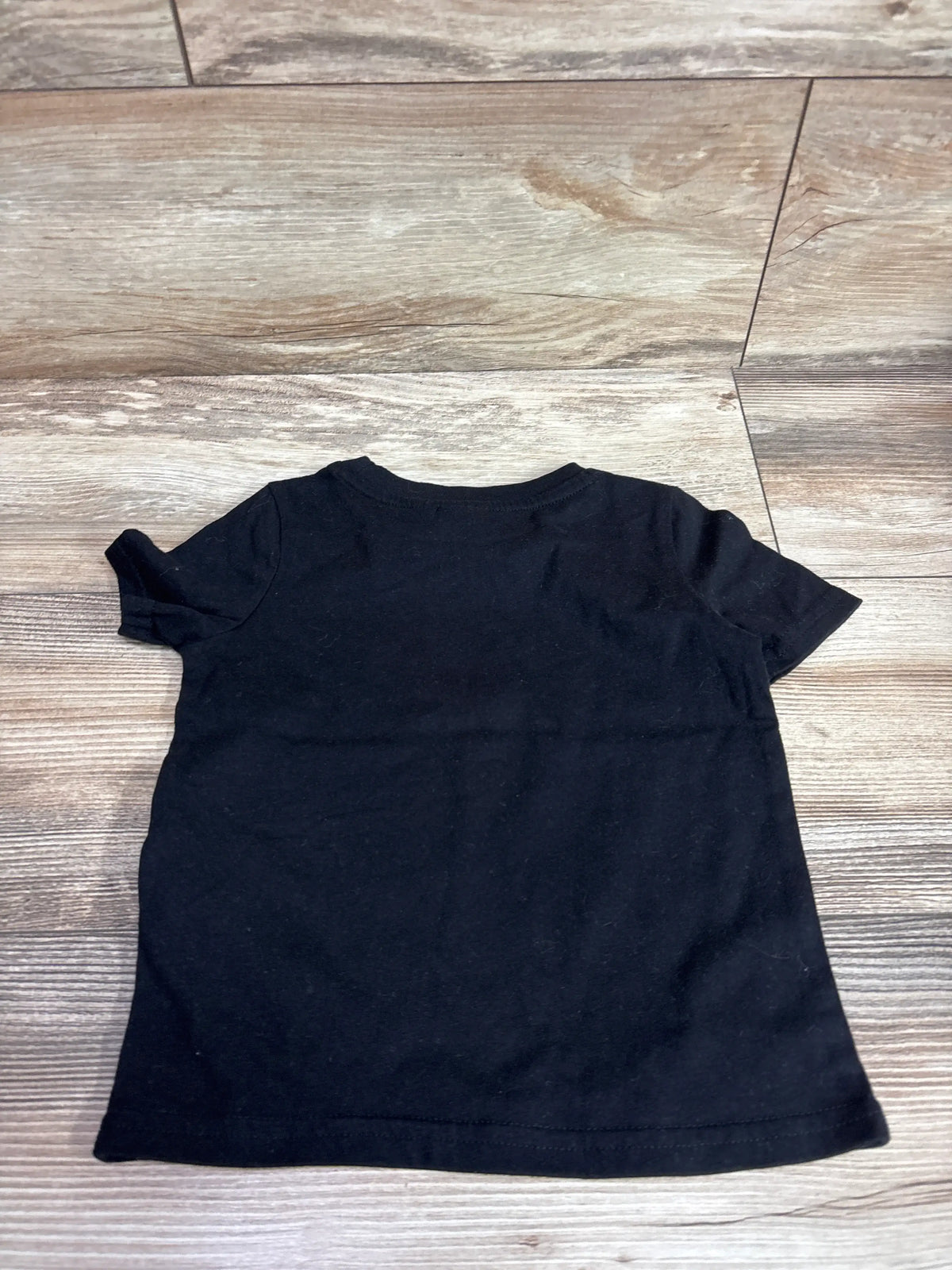 Back view of a plain black short sleeve t-shirt for a toddler size 2T on a wooden floor.