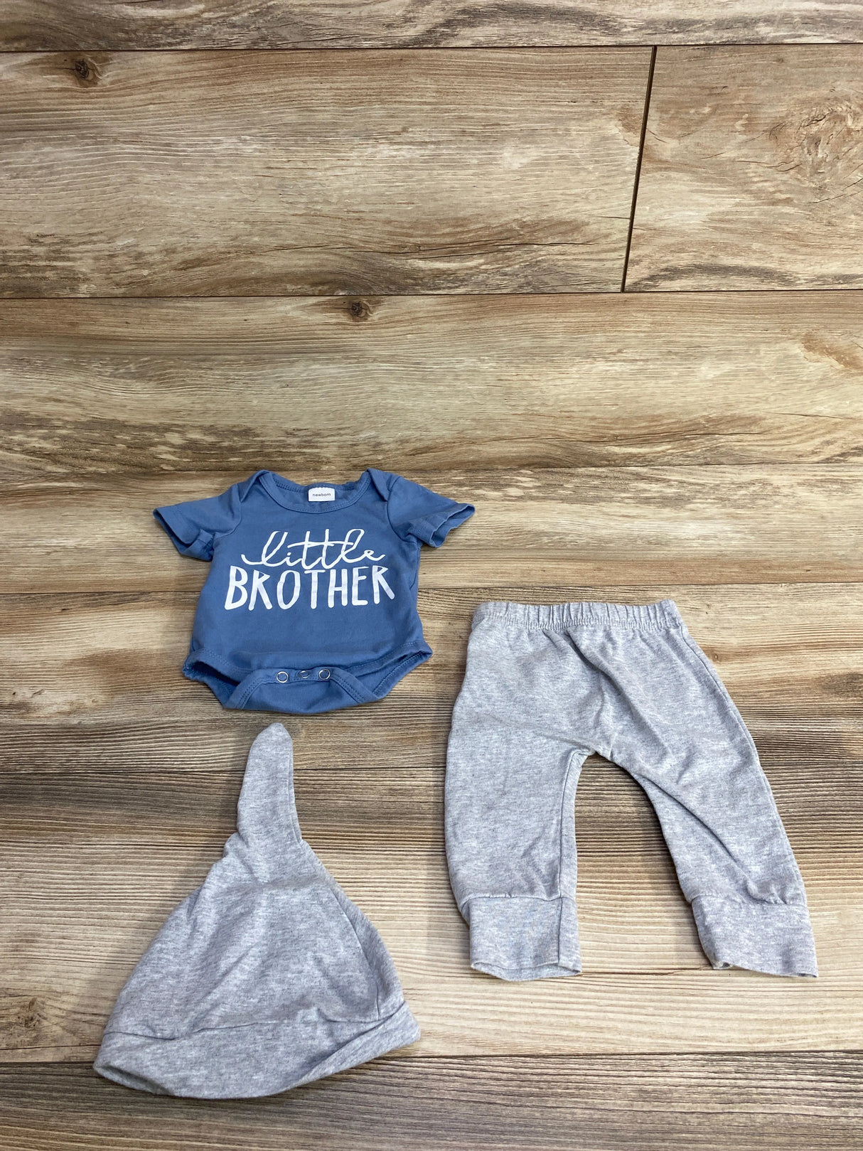 Newborn baby outfit set with blue "little brother" onesie, gray hat, and gray pants on wooden floor