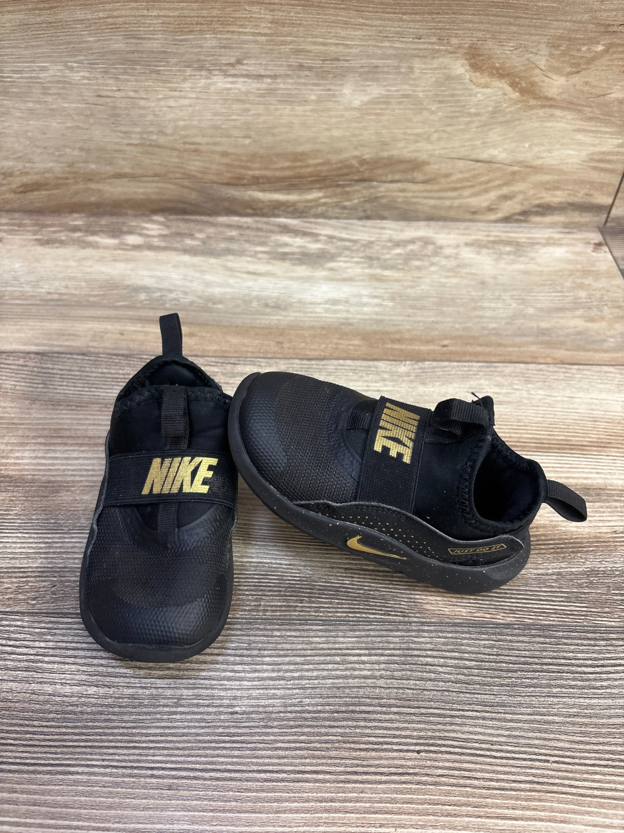 Side and front views of Nike Flex Runner toddler sneakers in black with gold NIKE strap and heel pull tabs on wood background