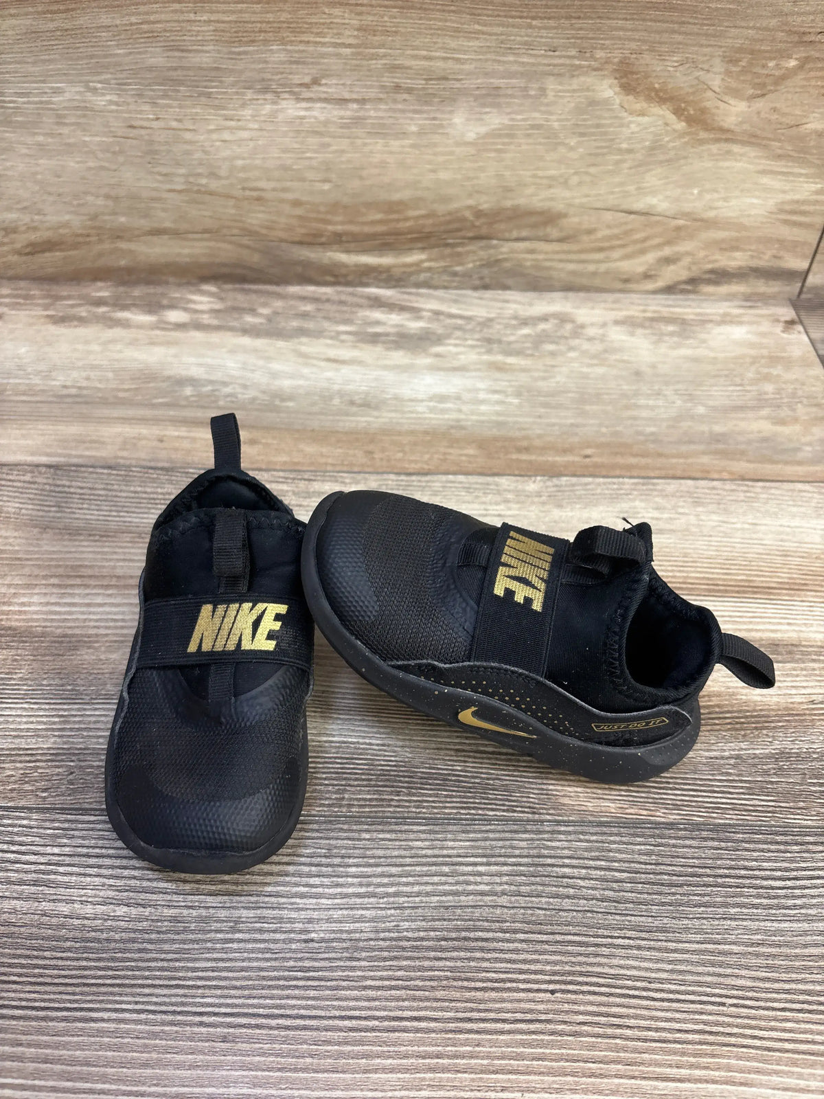 Side and front views of Nike Flex Runner toddler sneakers in black with gold NIKE strap and heel pull tabs on wood background