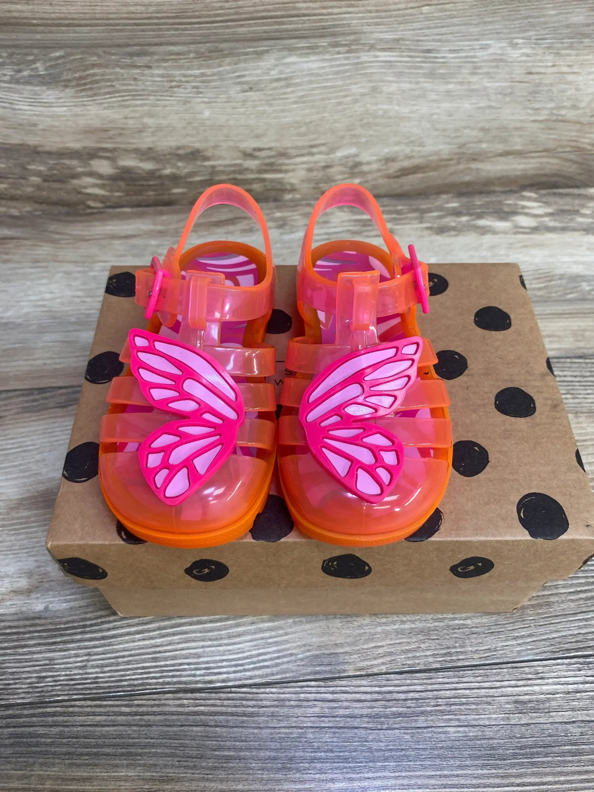 Top-down view of Sophia Webster toddler jelly sandals showcasing pink butterfly appliqués and translucent orange straps on a brown polka-dot box