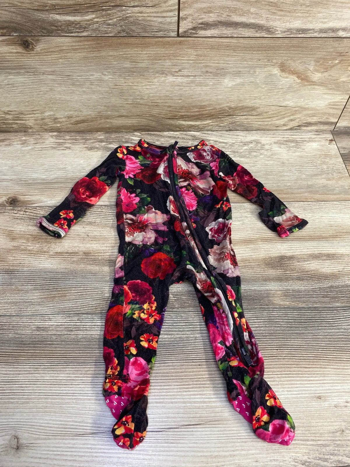 Front flat-lay of black floral baby footie by Alice + Olivia x Posh P showing diagonal zipper and footed legs on light wood background