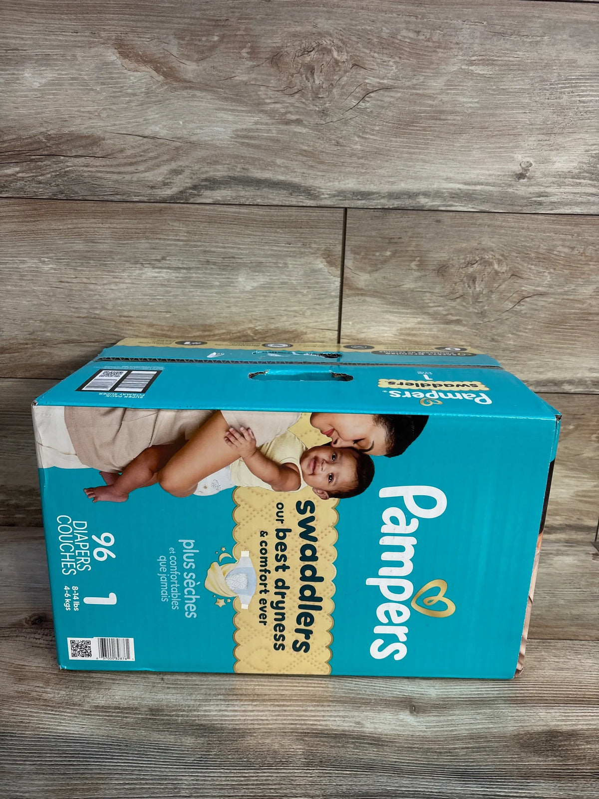 Front view of Pampers Swaddlers diapers Size 1 96-count box showcasing dryness and comfort claims on a wood floor