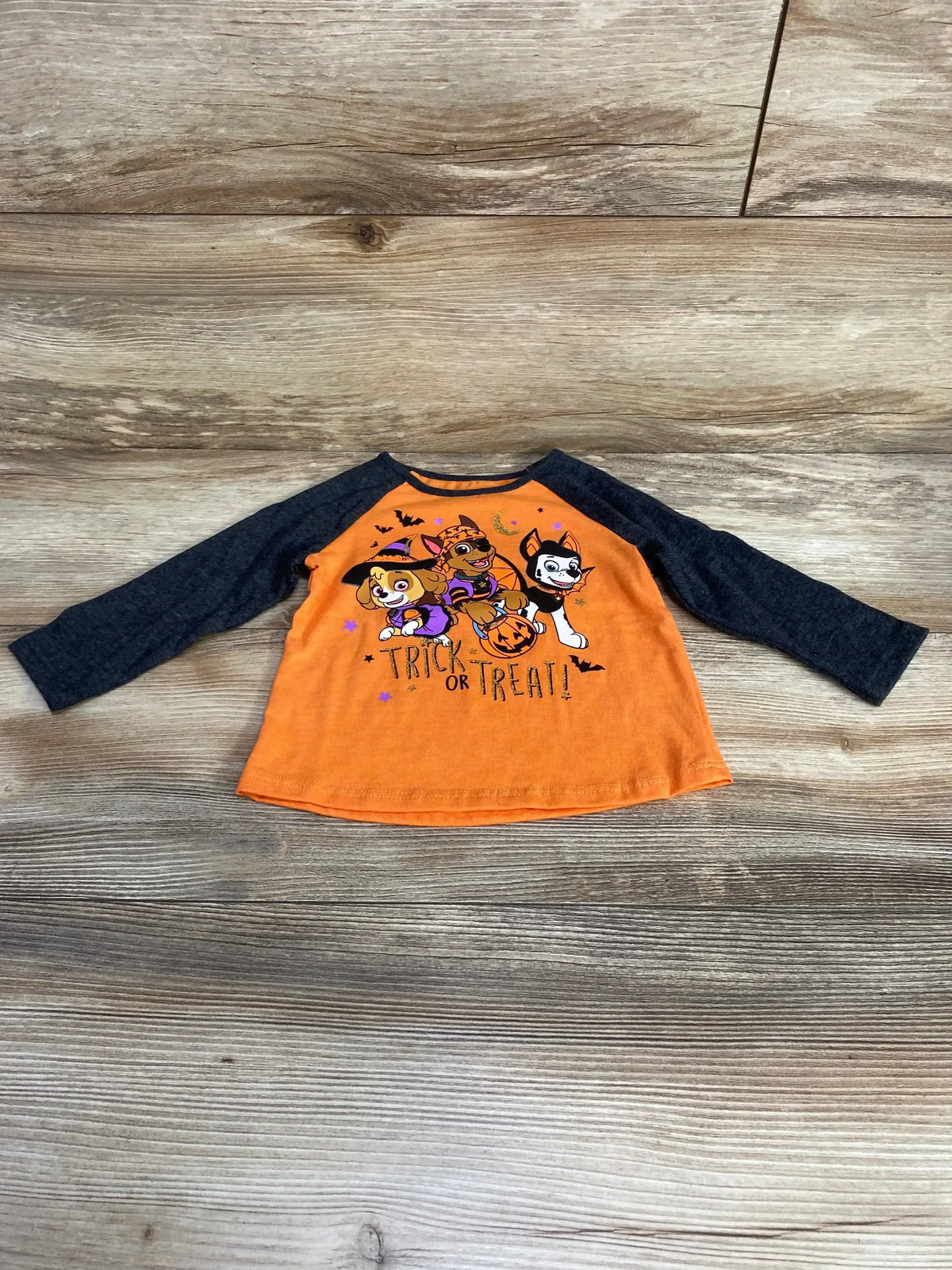 Front view of Jumping Beans Paw Patrol shirt with orange body, dark gray sleeves, and trick-or-treat graphic on wood