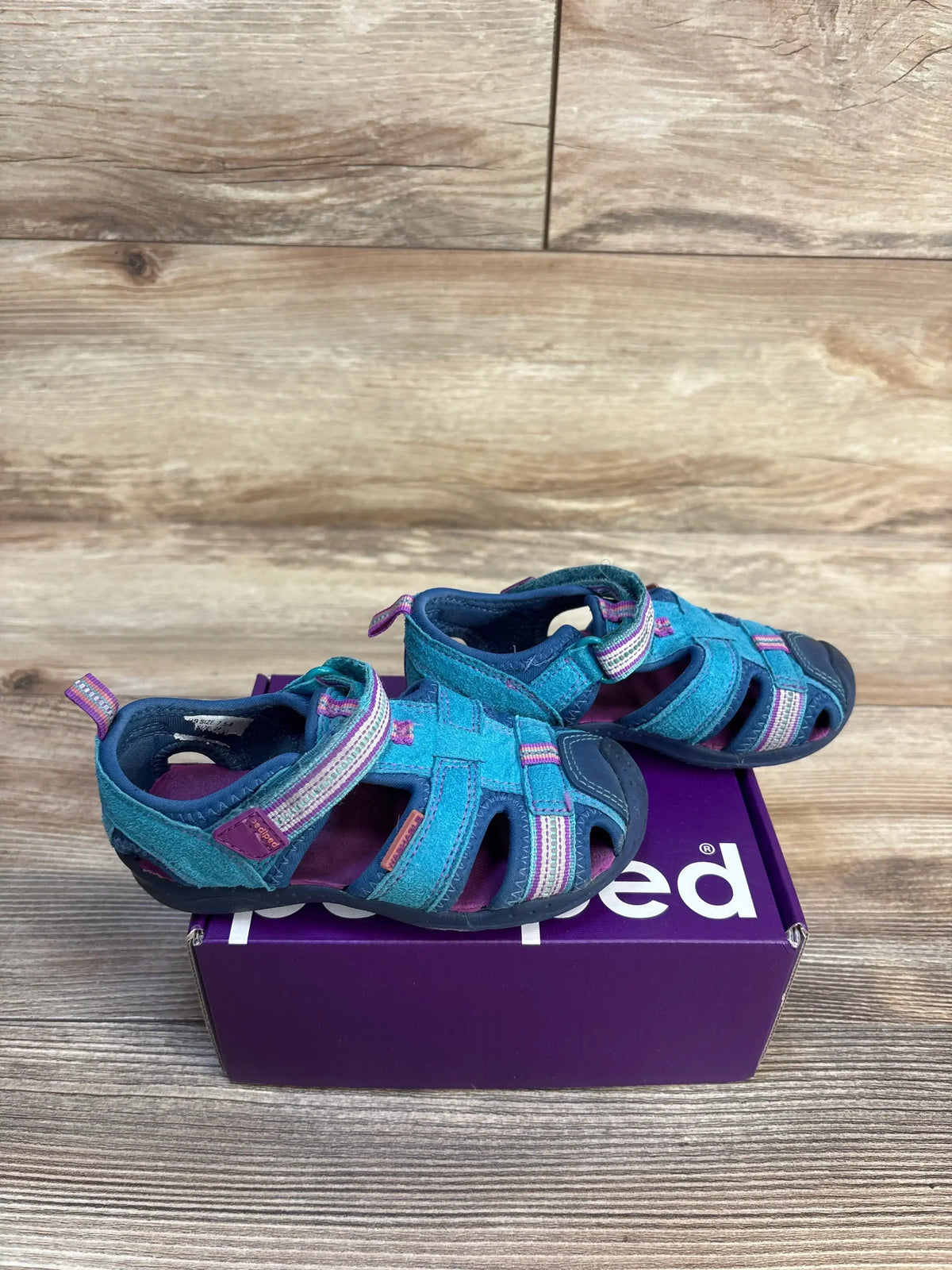 Teal and purple toddler sandals with velcro straps on a purple shoebox, wooden background