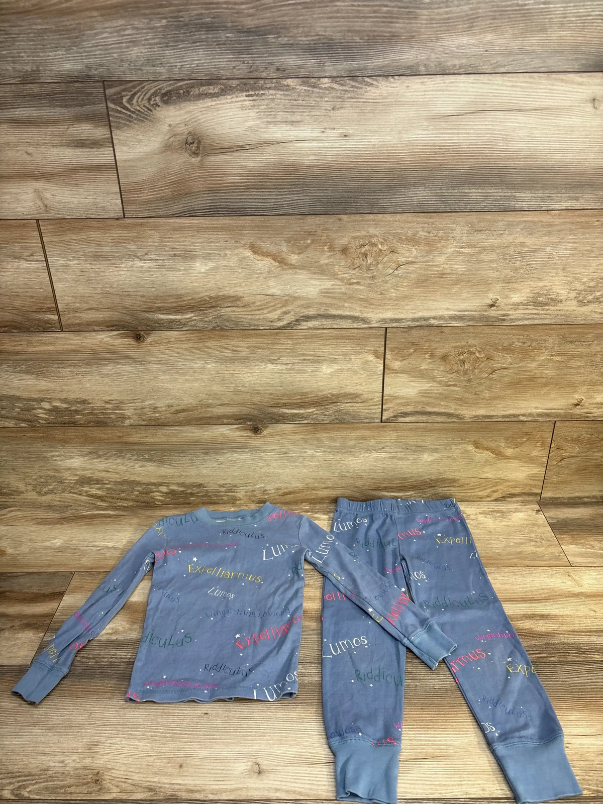 Flat lay of Mini Boden Harry Potter 2pc pajama set showing blue top and pants with colorful spell words on wood background