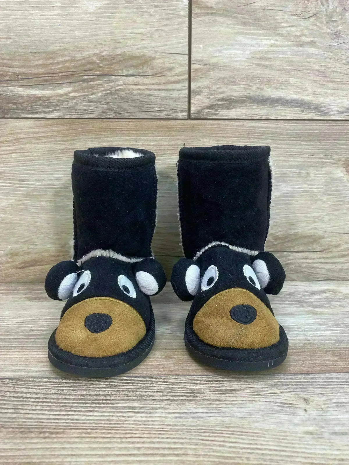 Front view of Lazy One kids’ black moose slipper boots showing plush moose face with tan snout and white eyes on wood floor
