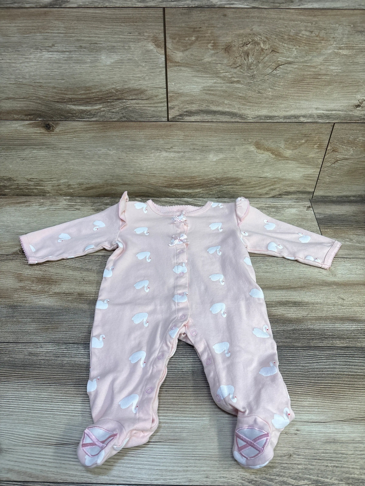 Front view of a pink swan print snap sleeper showcasing long sleeves, footed legs, and decorative bows on a wood grain background