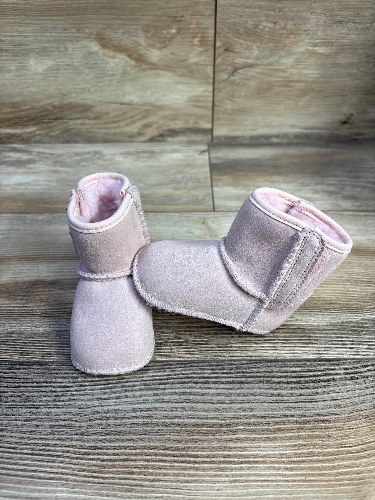 Front and side view of UGG Baby Classic Soft Sole boots in pink showcasing suede exterior and plush shearling lining on wood backdrop