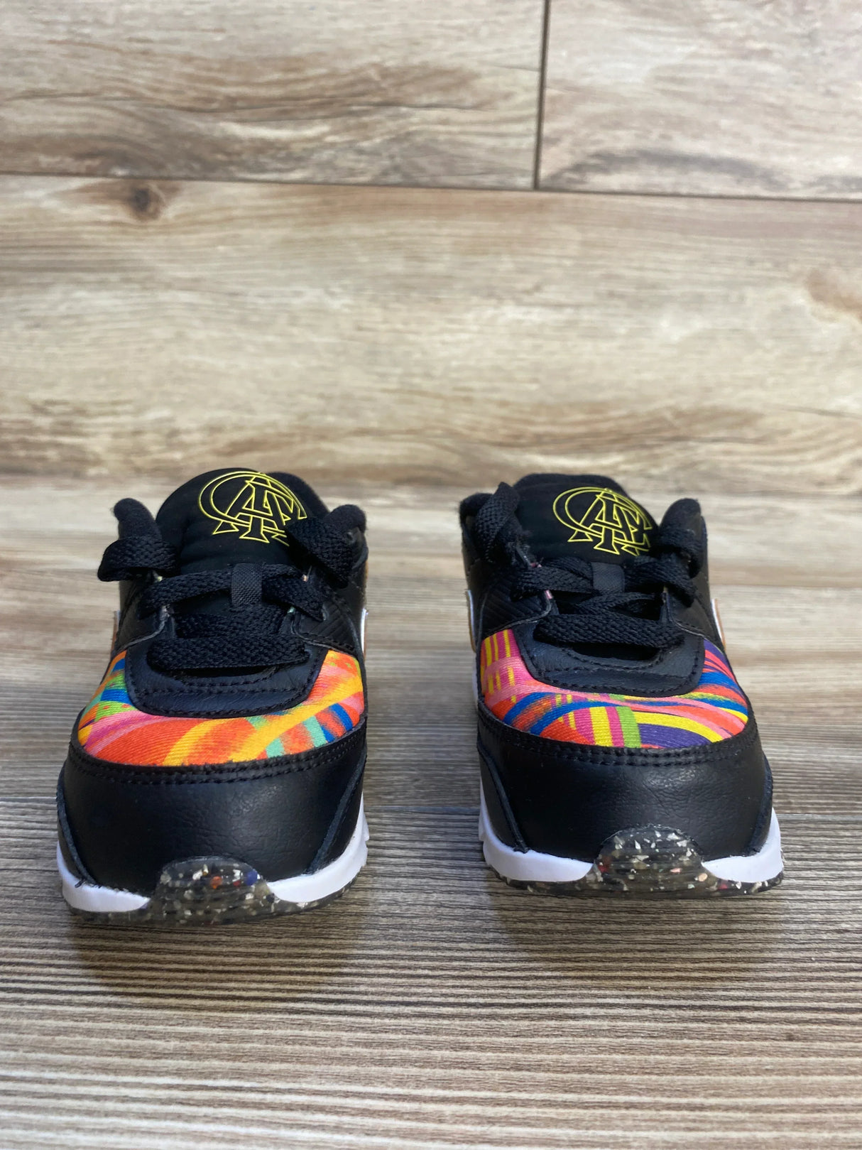 Front view of Nike Air Max 90 TD 'Latino Heritage Month - Familia' kids sneakers, black with multicolor patterned toe and white sole on wood floor