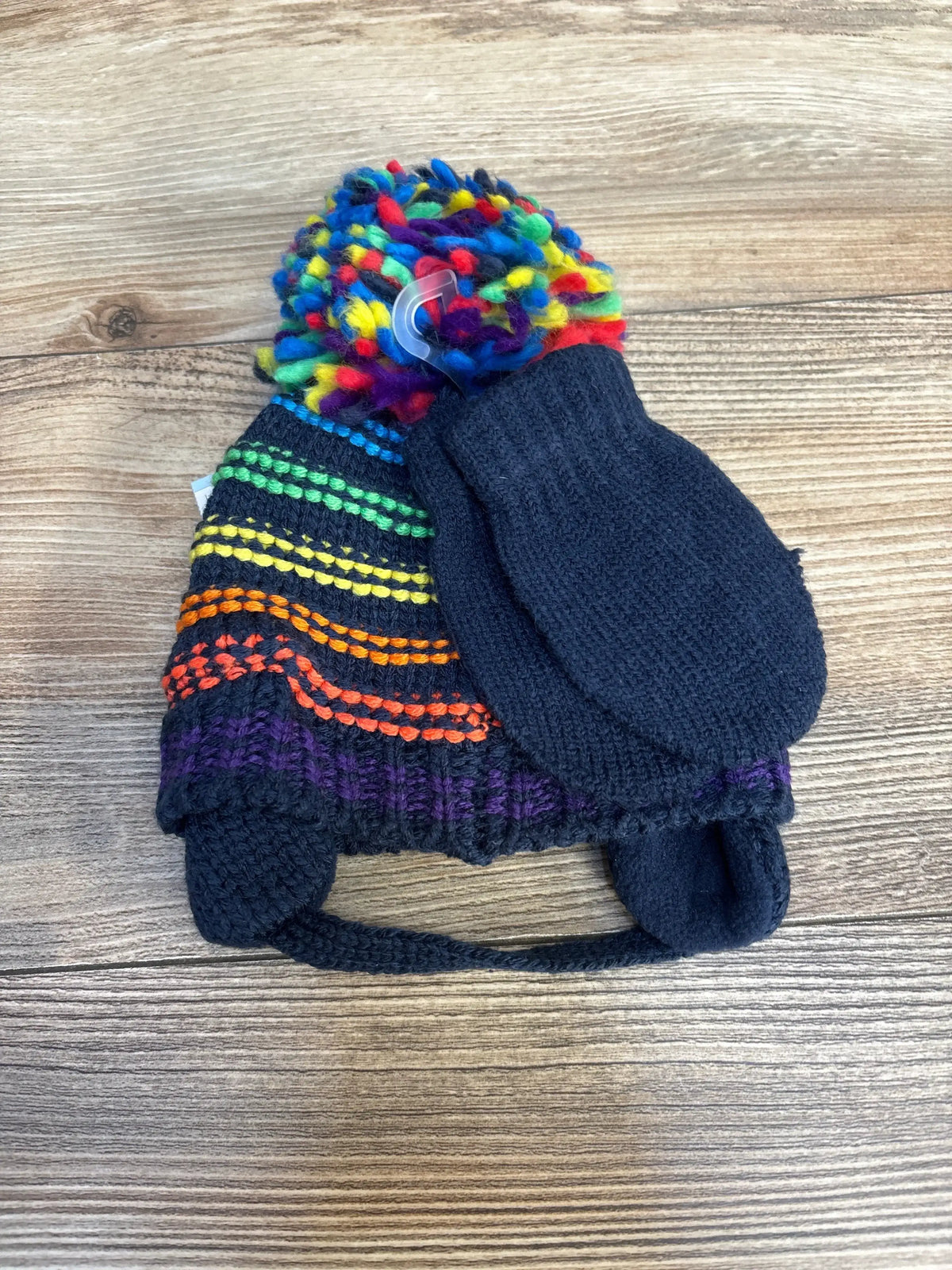 Knit kids winter hat with rainbow pom pom and dark knit mittens on wood background