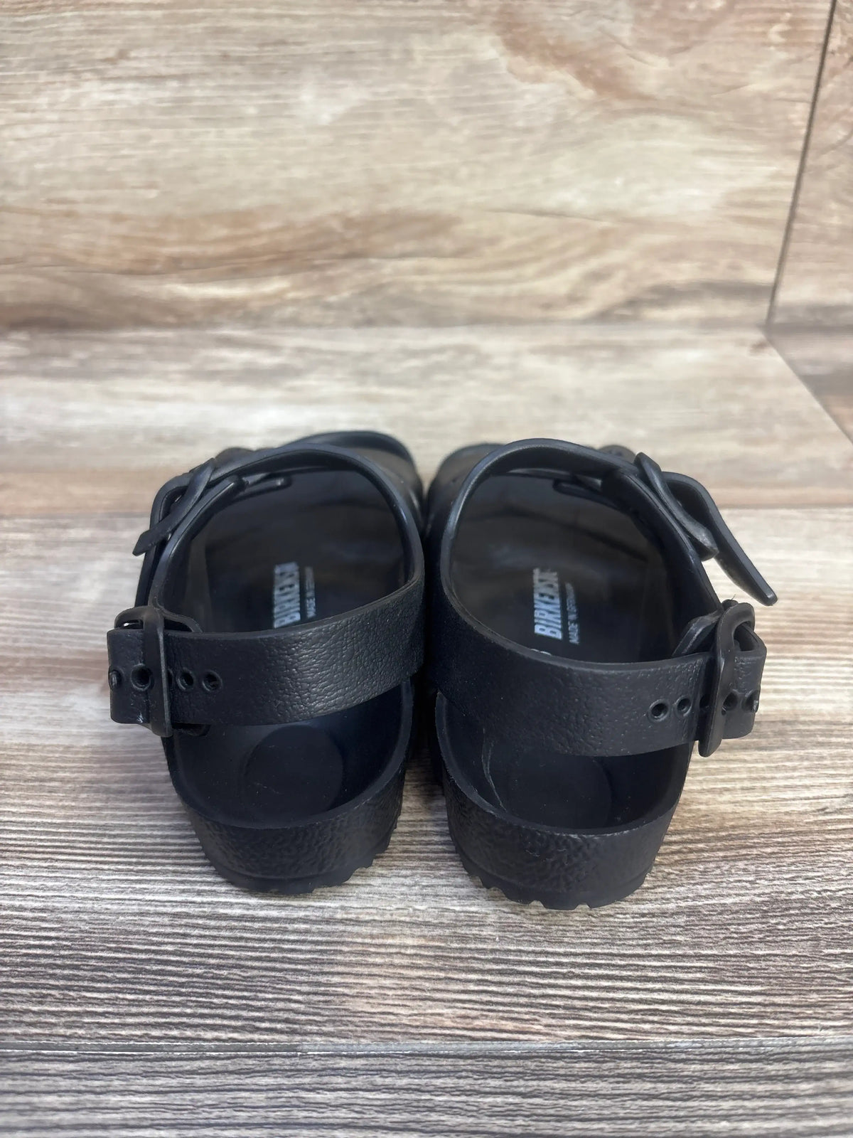 Front view of black Birkenstock Milano EVA sandals in kids size 8C showcasing adjustable buckled straps on a wood floor
