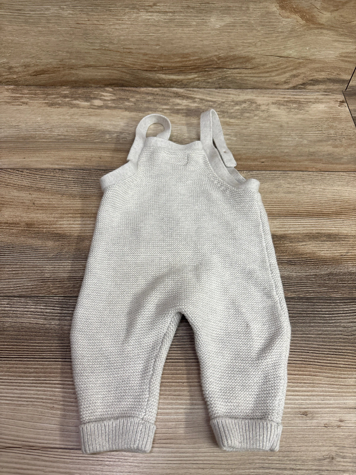 Flat lay of grey Little Planet knit overalls, size 3 months, with shoulder straps and ribbed cuffs on a wood floor