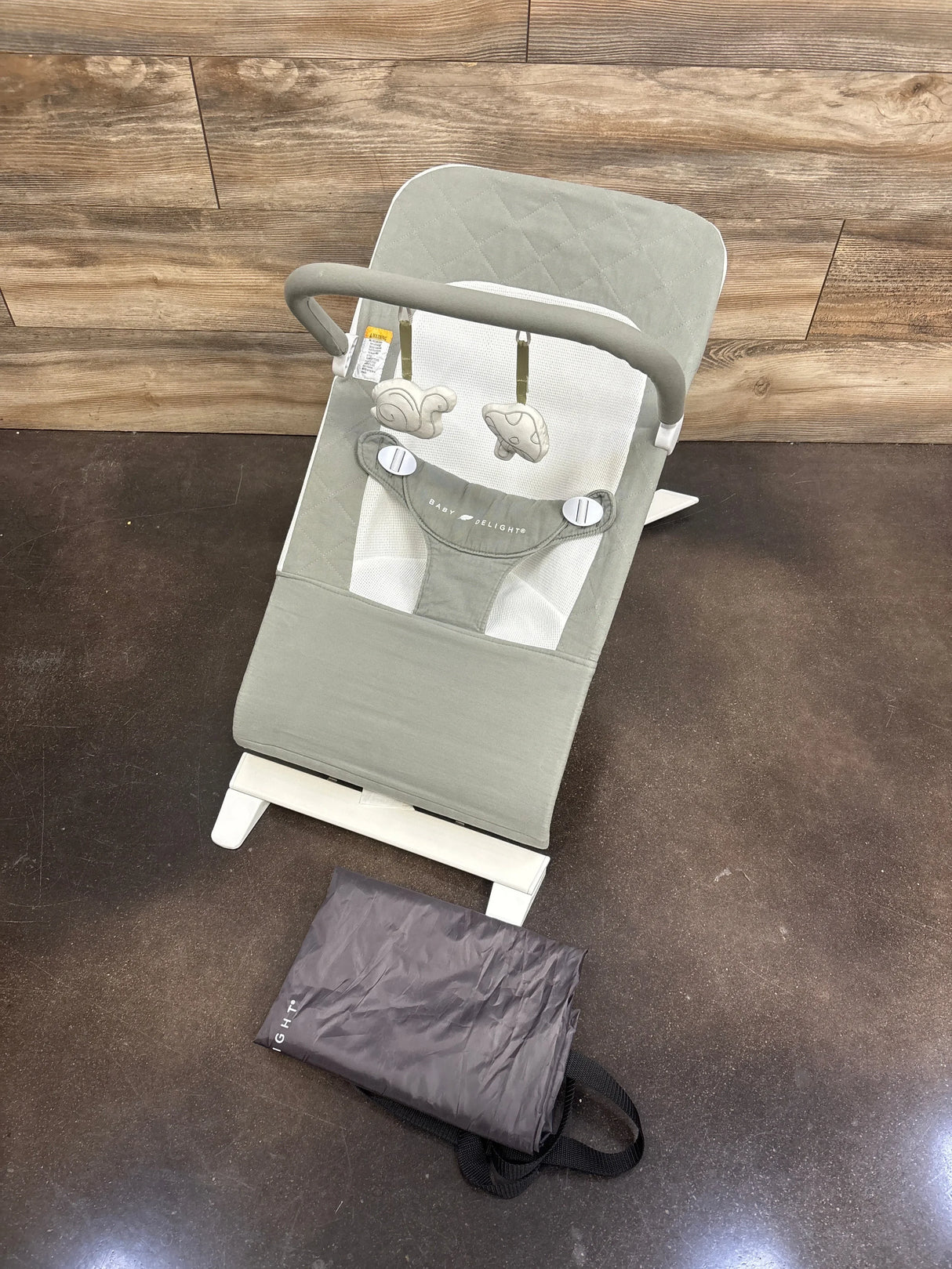 Overhead view of Baby Delight Alpine Deluxe bouncer in sage showing mesh seat and toy bar with two hanging toys on dark floor