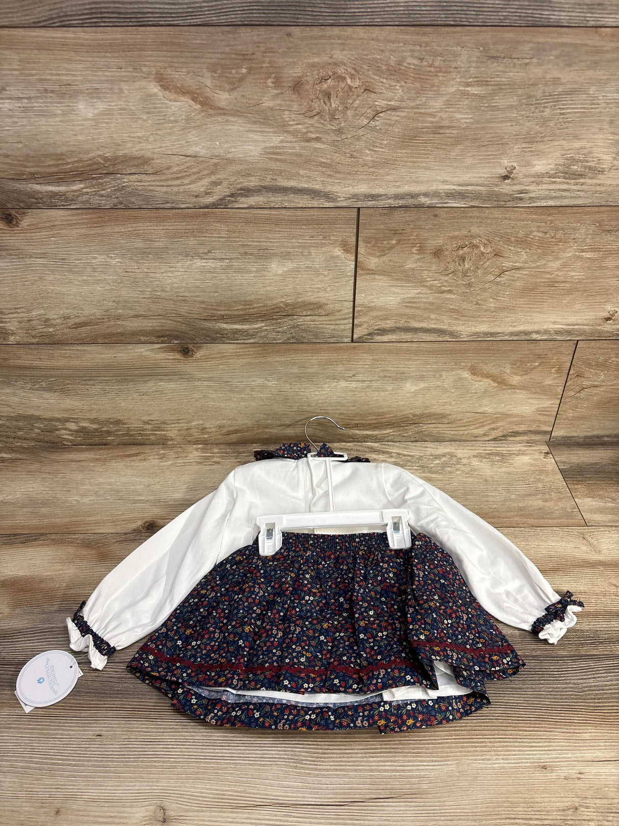 Flat lay of Edgehill Collection white long-sleeve bodysuit and navy floral skirt set showing ruffle collar and elastic waist on wood floor
