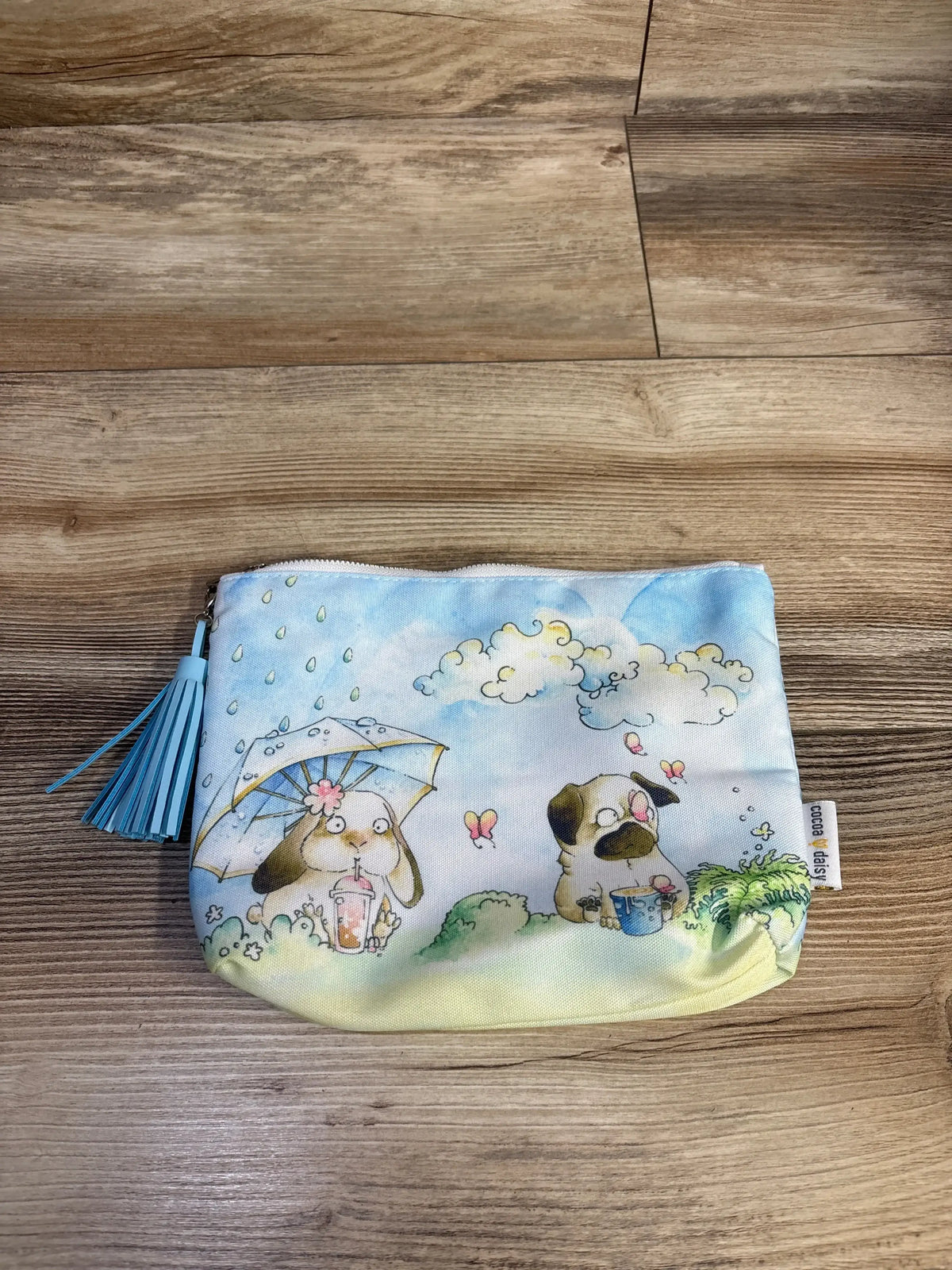 Flat lay of Cocoa Daisy wet/dry snack bag showing puppy and bunny illustrations and blue tassel zipper on wood background