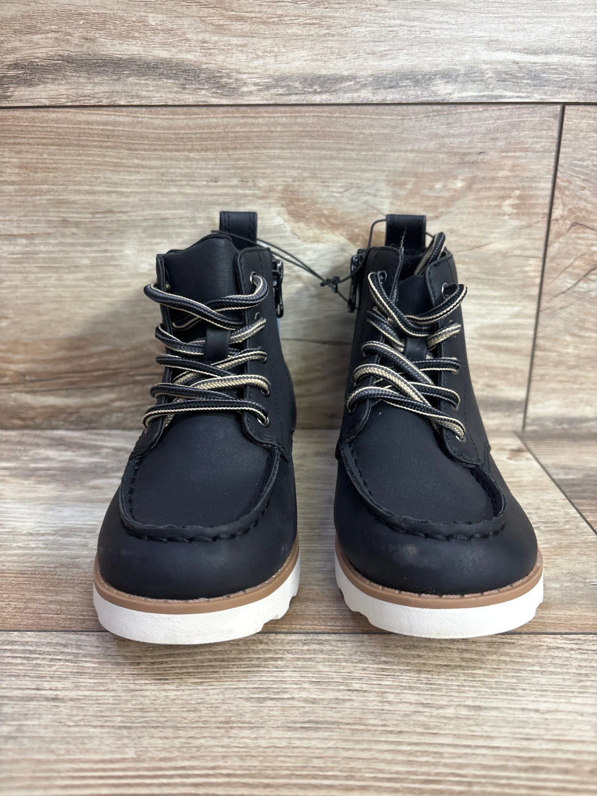 Front view of kids black lace-up boots showcasing matte black uppers with white striped laces and white rubber soles on wood plank floor