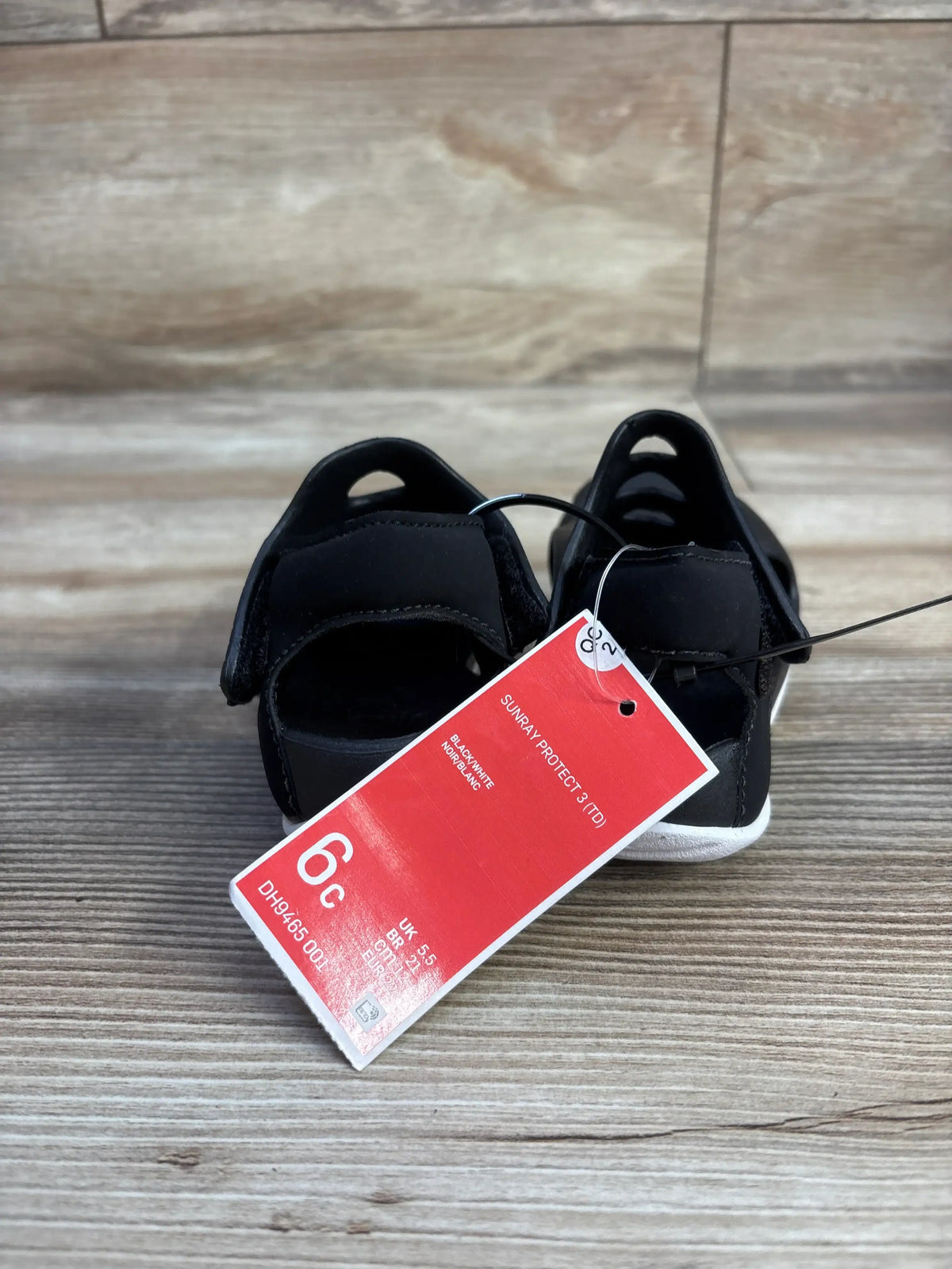 Front view of black Nike Sunray Protect 3 sandals showing open toes, adjustable straps, and a red size tag on wood-look flooring