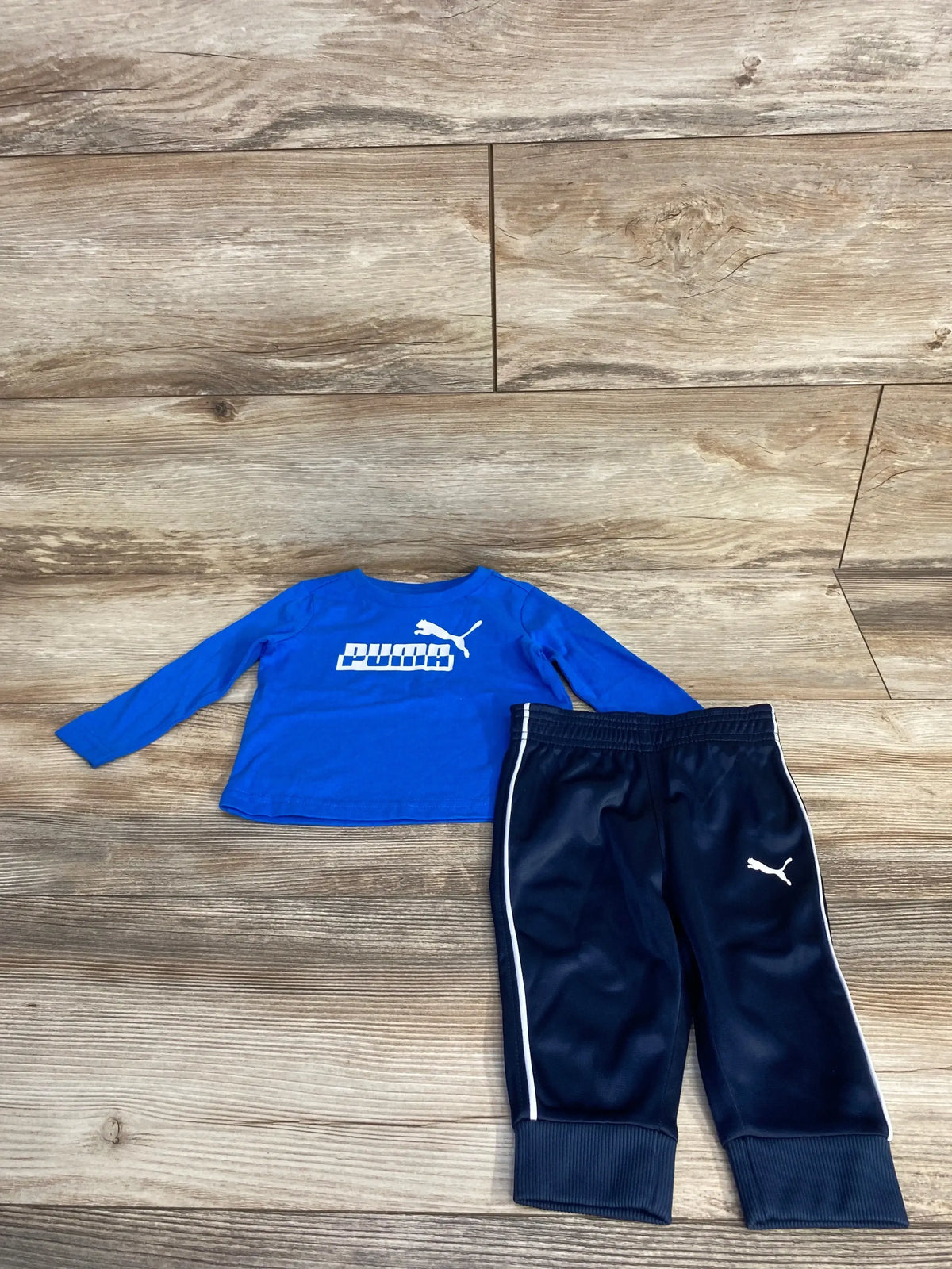Flat lay of blue Puma long-sleeve logo shirt and navy Puma jogger pants showing white Puma wordmark and piping on wood floor