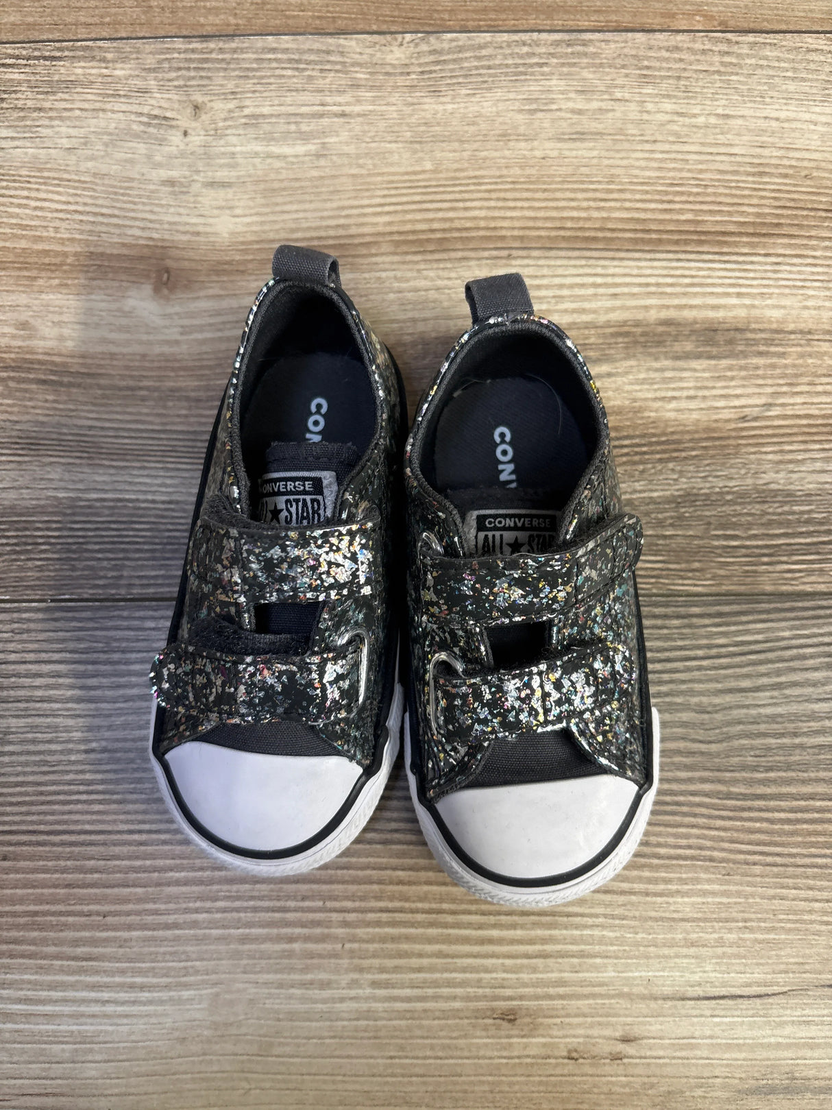 Top view of toddler Converse size 6C glittery black sneakers with Velcro straps and white toe caps on wood floor