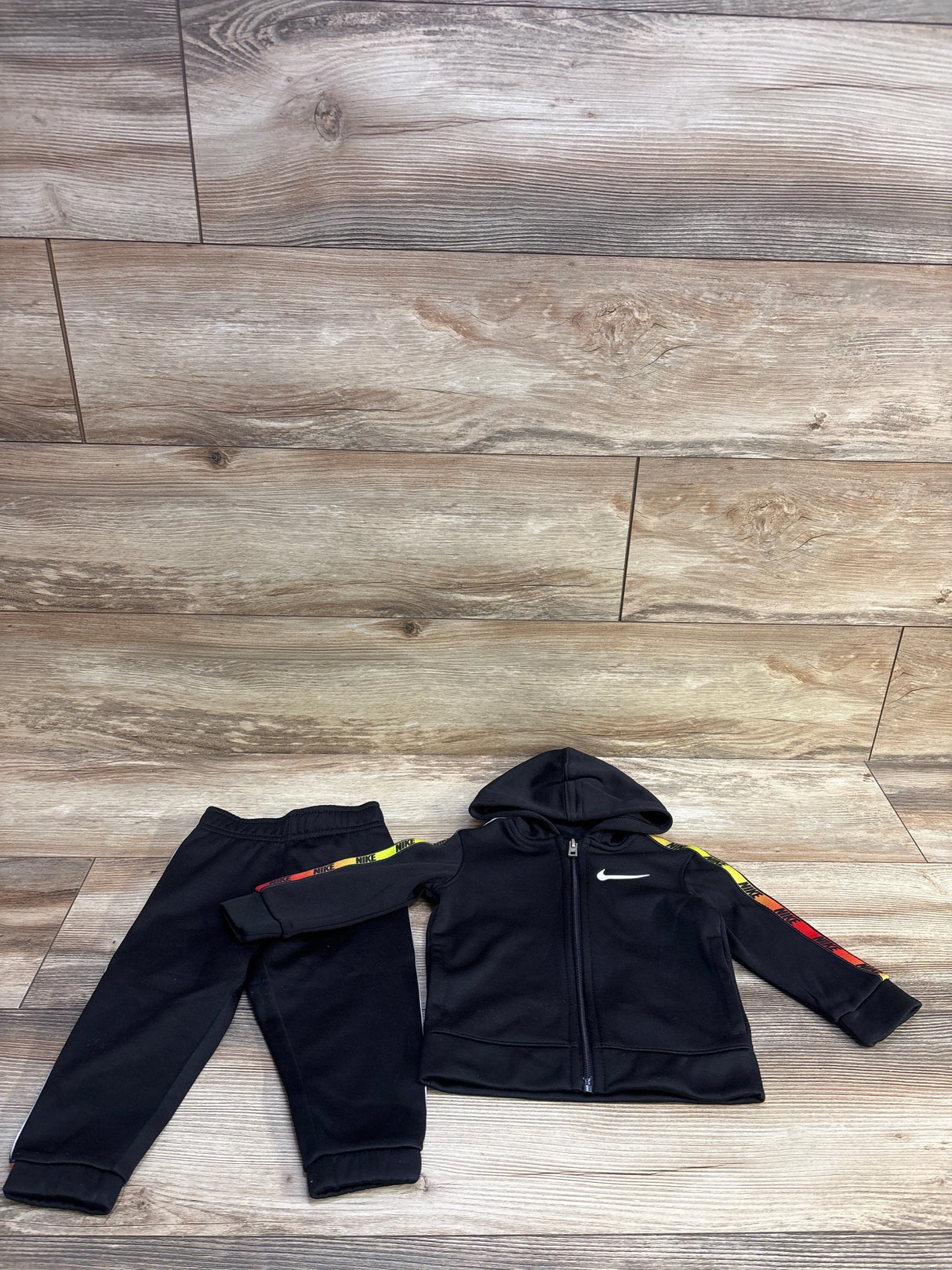Flat lay of black Nike 2-piece tracksuit size 12M, zip hoodie with white Swoosh and rainbow sleeve stripe on wood floor