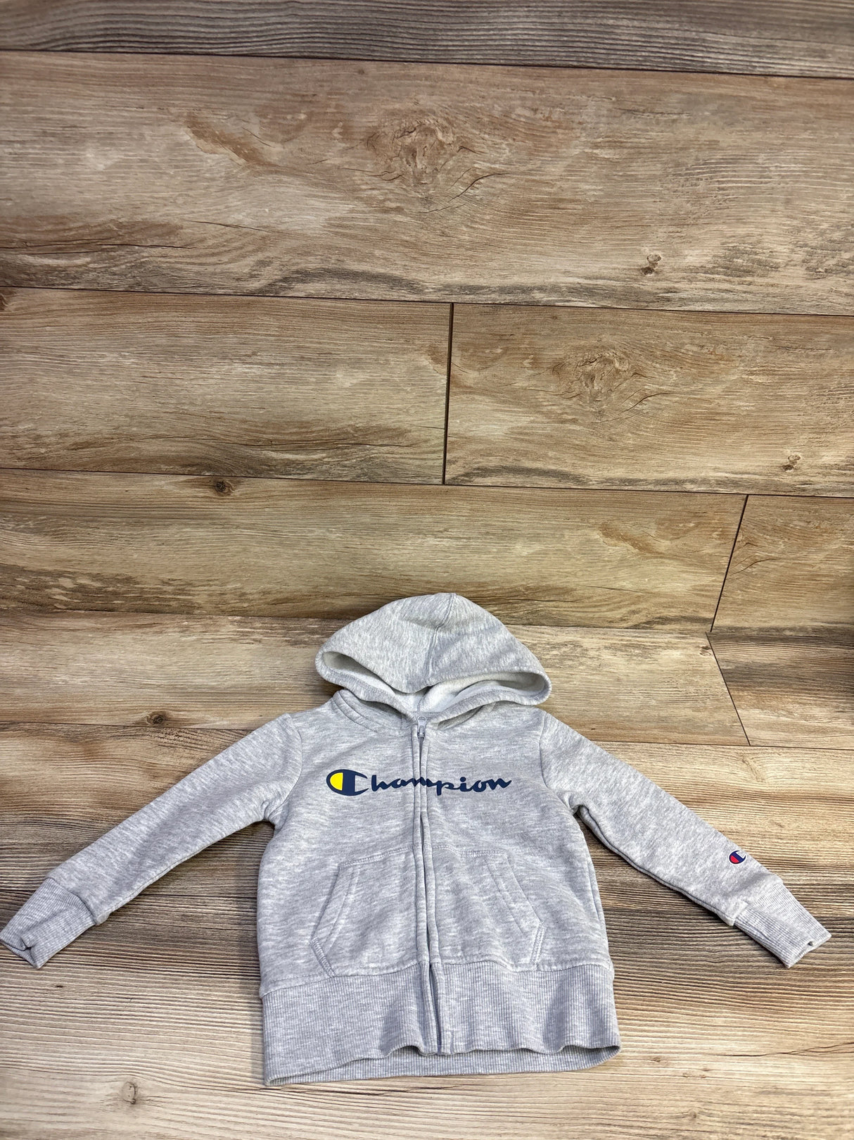 Flat lay of grey Champion toddler full-zip hoodie, size 2T, showing hood, front zip, kangaroo pocket and chest logo on wood floor