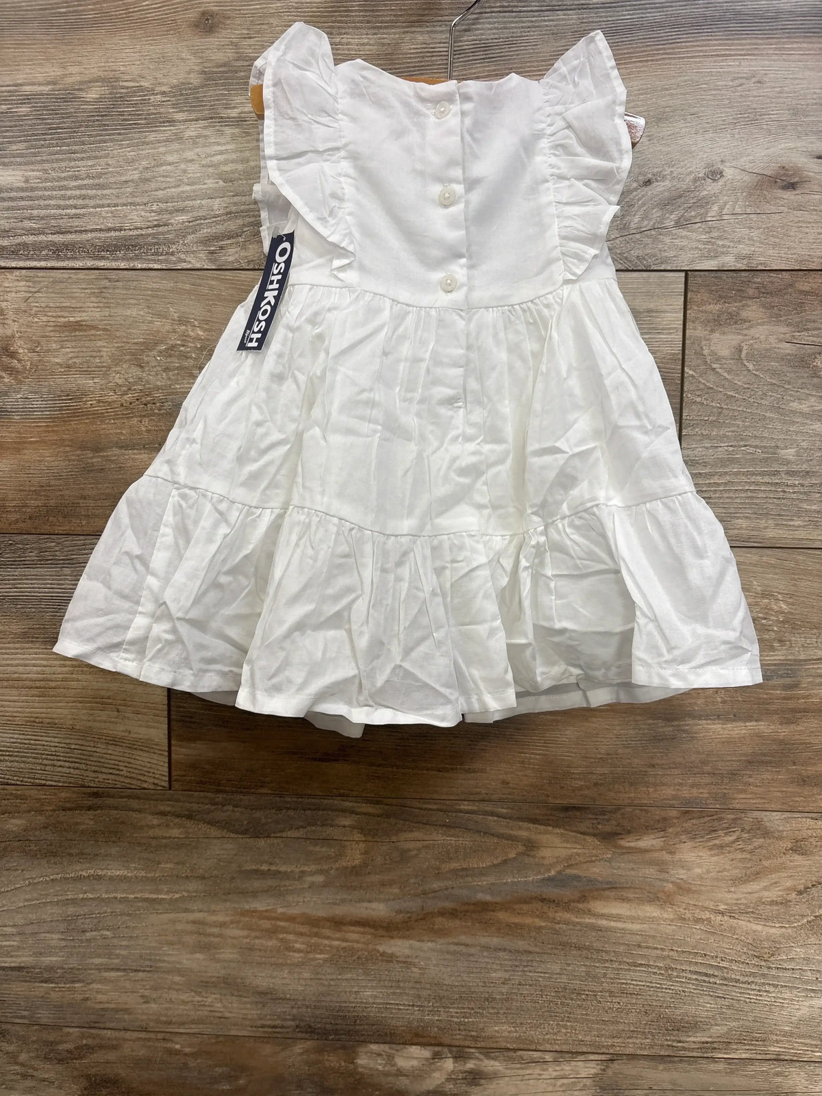 Top-down view of OshKosh B'gosh white toddler lace dress showcasing ruffled cap sleeves, button-back closure, and tiered skirt on wood floor background