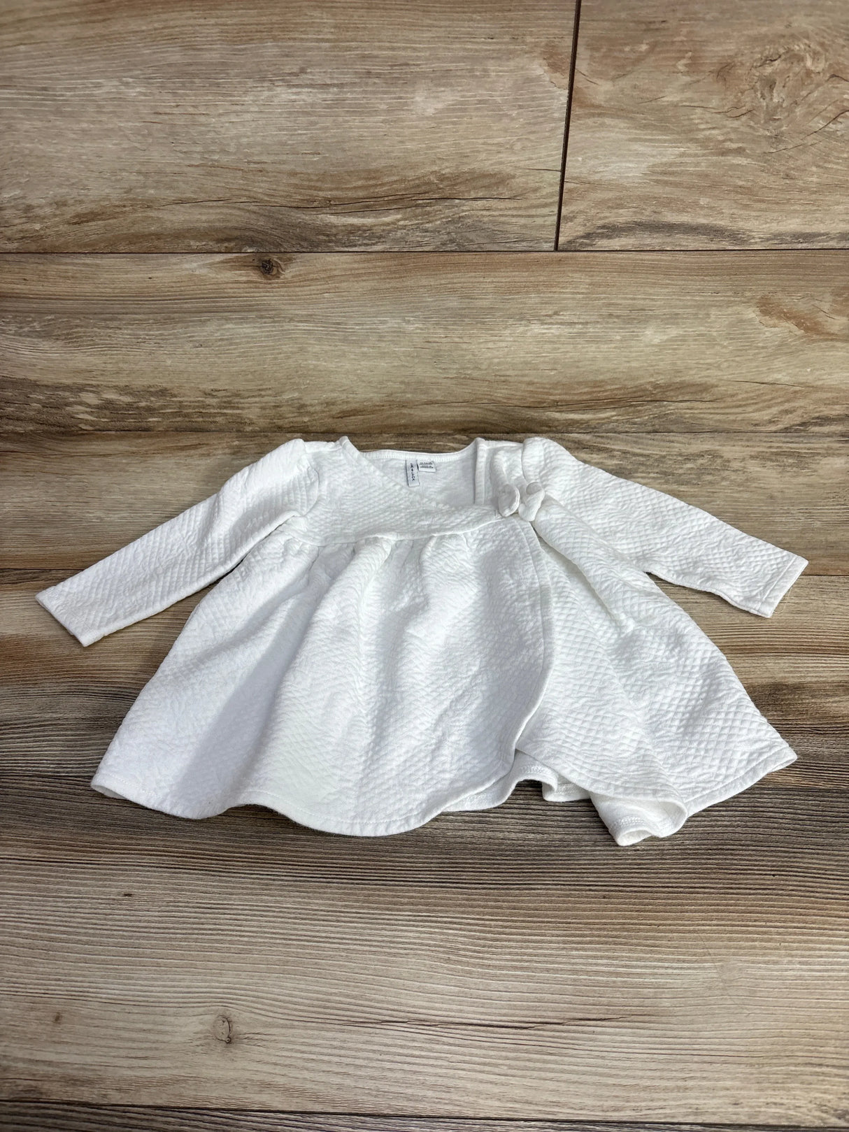 White textured long sleeve baby dress with bow detail, laid flat on wood background