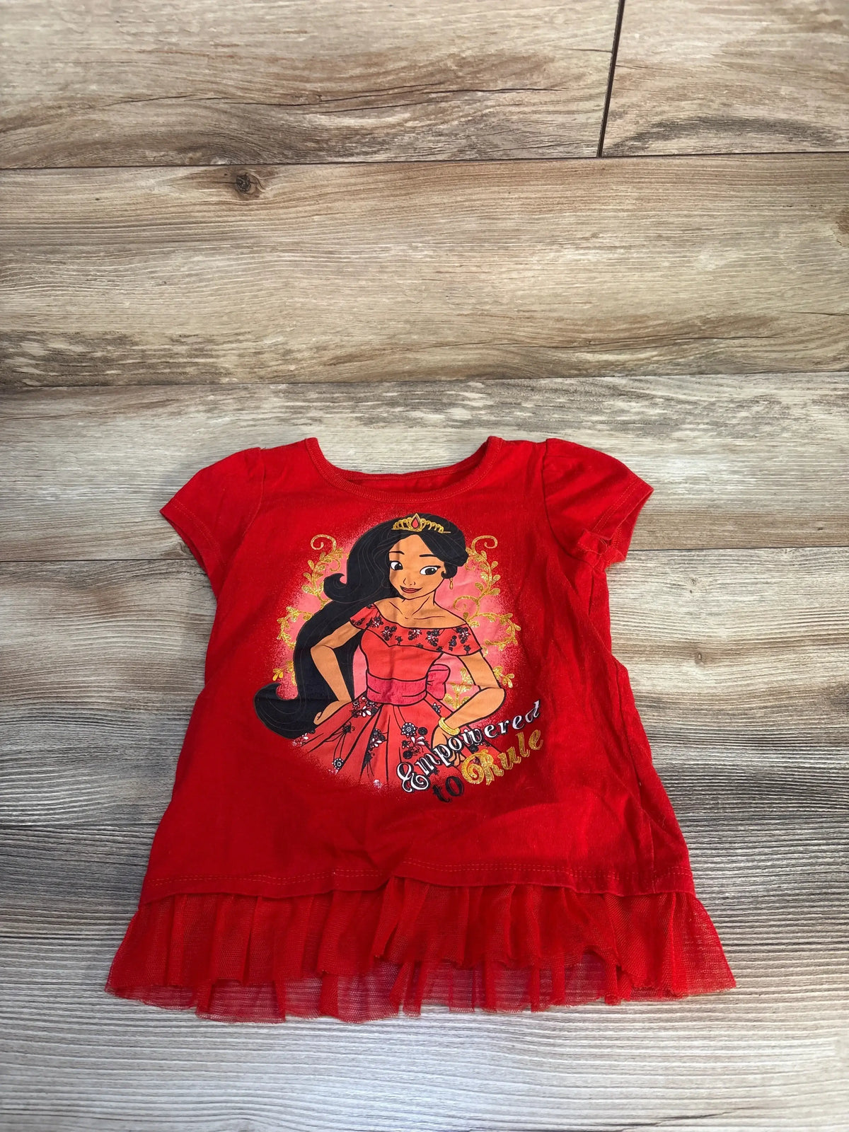 Front view of red Elena of Avalor peplum toddler shirt with tulle ruffle and Empowered to Rule graphic on wood floor