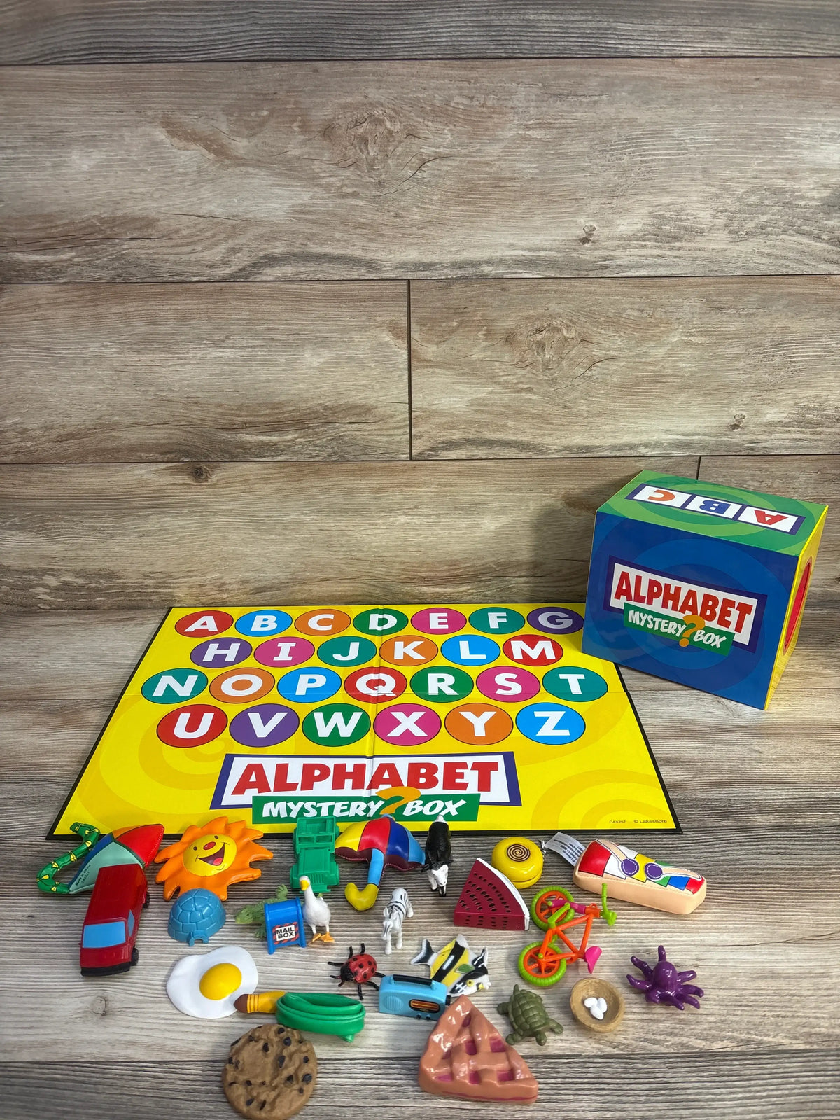 Angled view of Lakeshore Alphabet Mystery Box showcasing the uppercase letter game board and 26 miniature objects on a wood surface