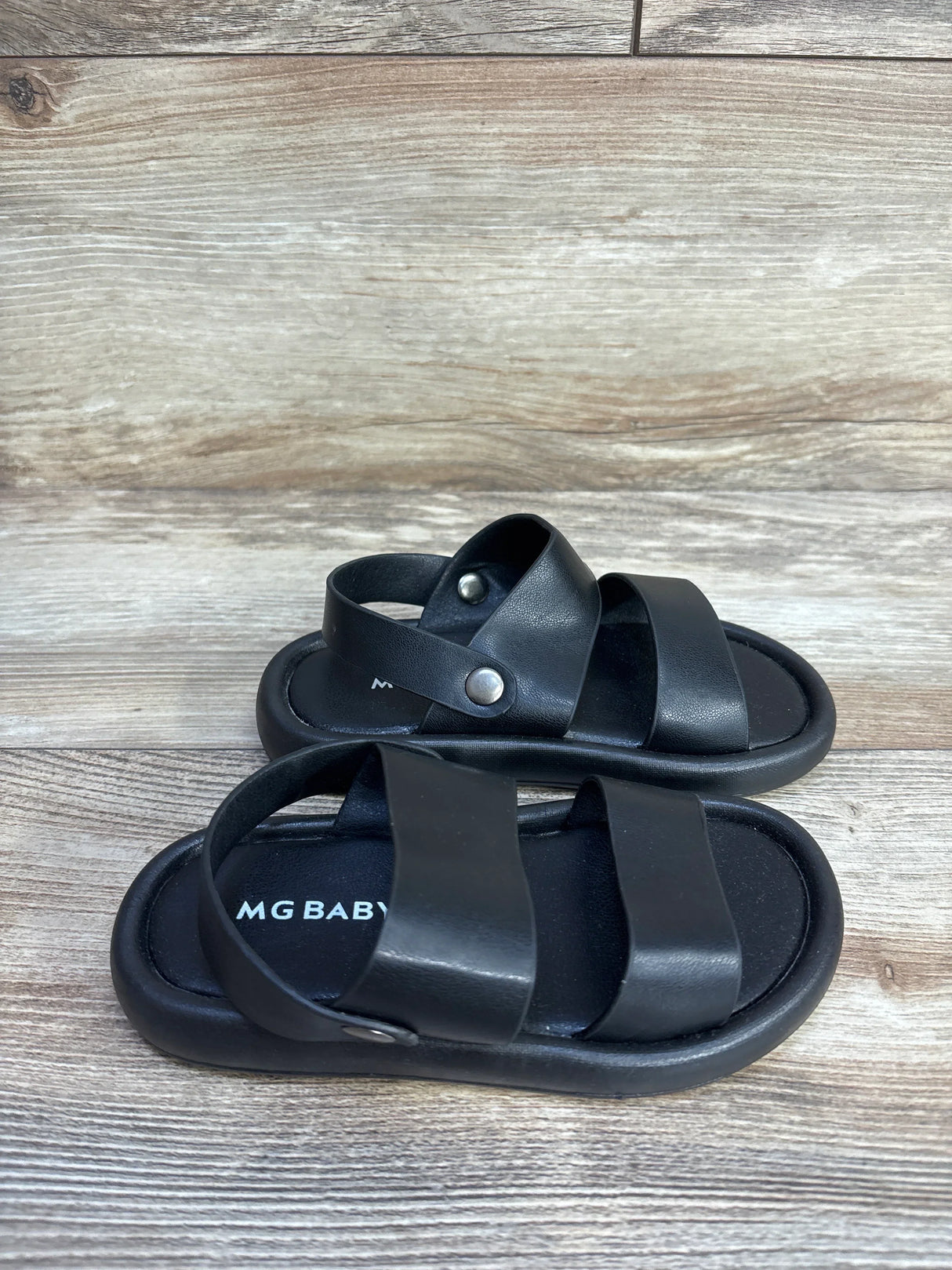 Three-quarter view of MGBABY black foam sandals size 11/12c showing two wide straps and snap heel strap on wood background