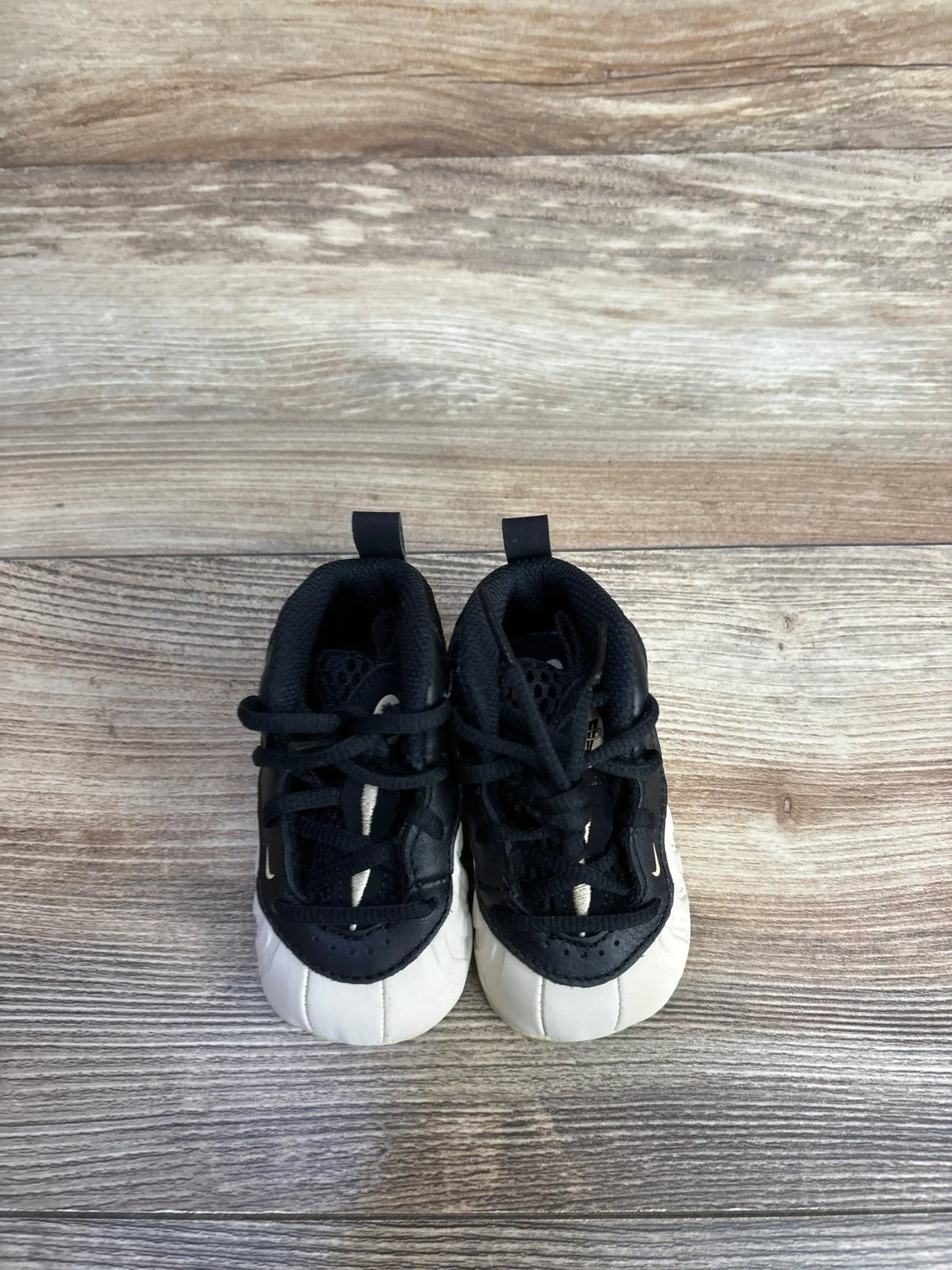 Top-down view of Nike Posite One crib booties (size 3C) showcasing black upper, white rounded toe caps, black laces and heel pull tabs on a wood floor