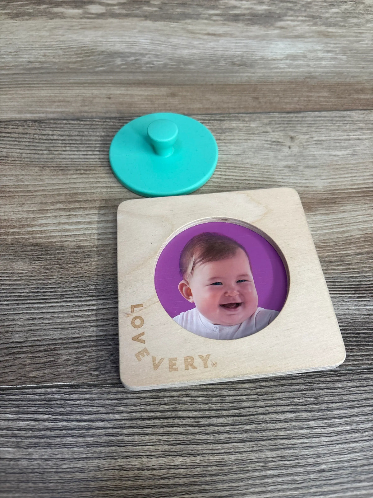 Top view of Lovevery First Puzzle wooden piece with circular baby photo insert, engraved LOVEVERY logo, and detached teal knob on wood surface