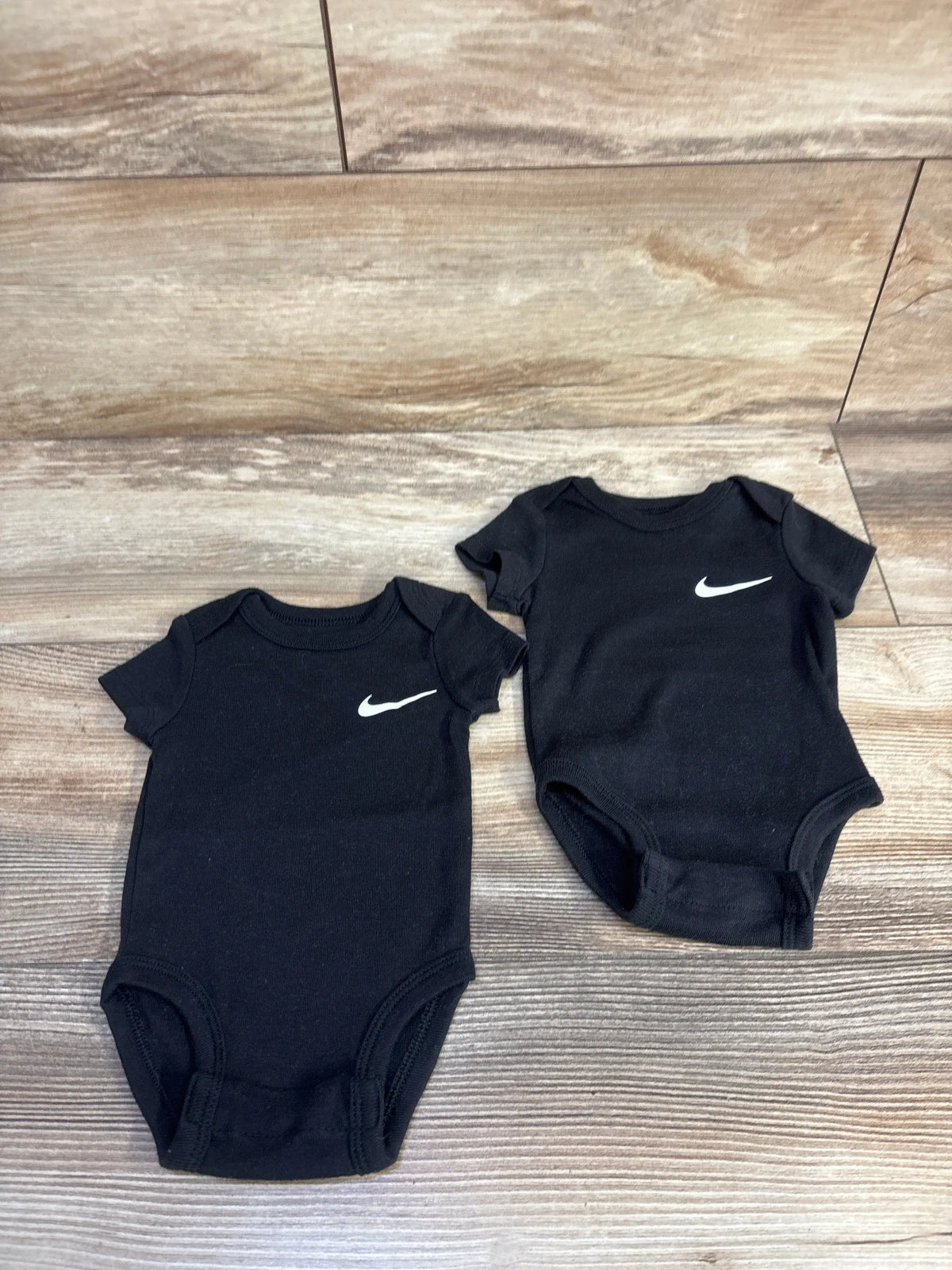 Top view of Nike 2pk bodysuits in black, size 3 months, showcasing short sleeves and swoosh logos on a wood floor