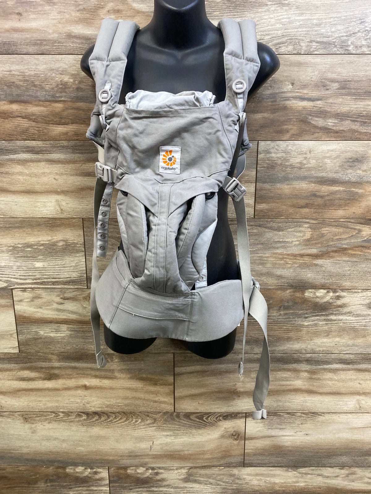 Front view of Ergobaby Omni 360 carrier in pearl grey with padded straps, lumbar belt, adjustable buckles on wood background