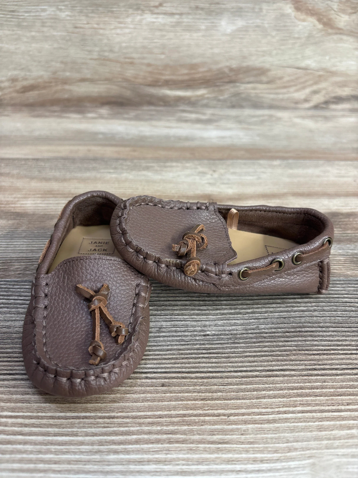 Angled front view of Janie & Jack brown leather driving shoes size 5C showing stitched moccasin toe, lace bow and pebbled leather on a wood surface