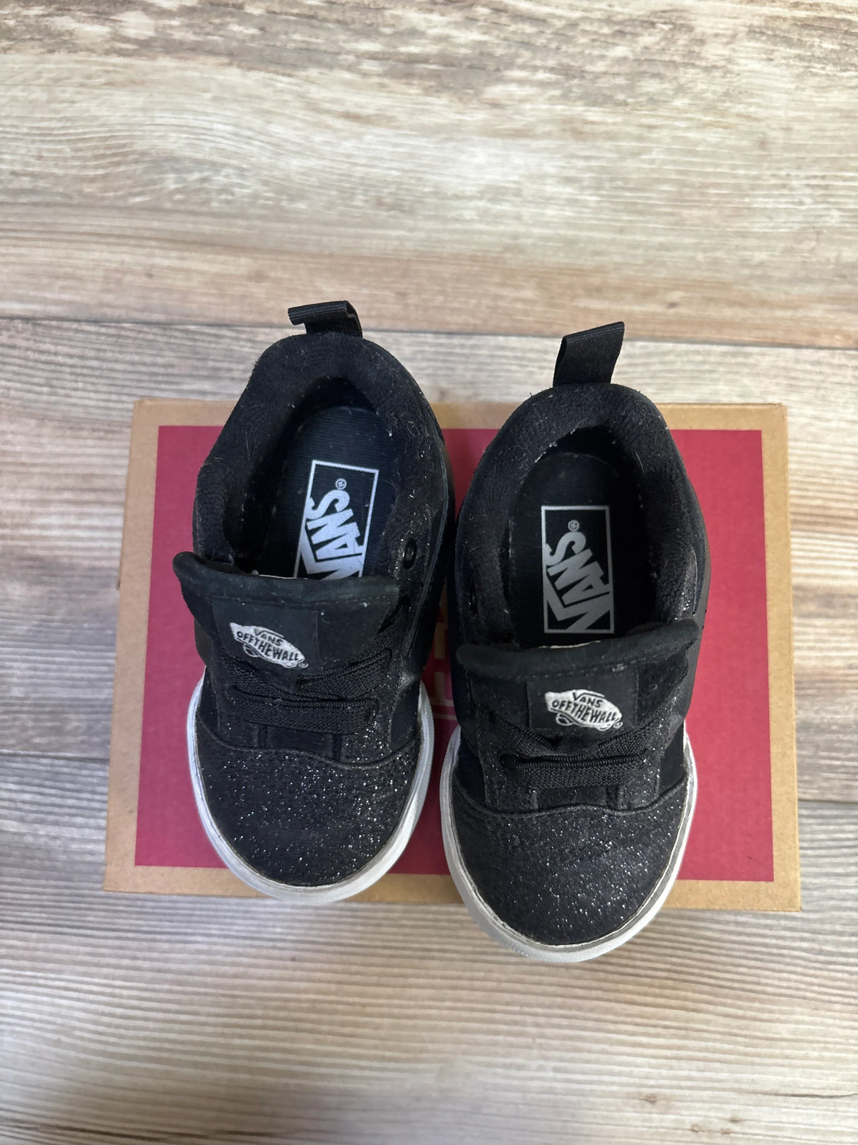 Top-down view of Vans Knu Skool toddler sneakers showcasing black glittered toes and white rubber soles on red shoebox