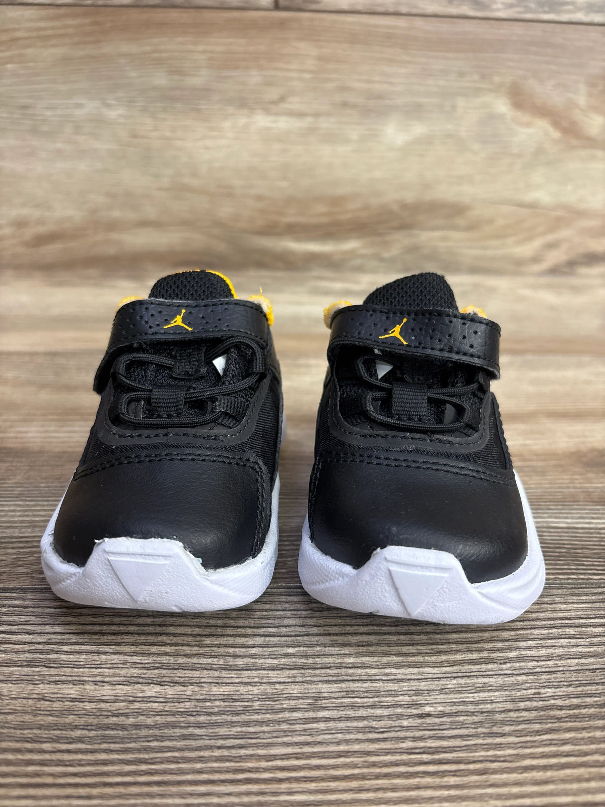 Front view of toddler Air Jordan 11 CMFT Low TD ‘Black Taxi’ sneakers with Velcro strap, yellow Jumpman logo, and white soles on wood background
