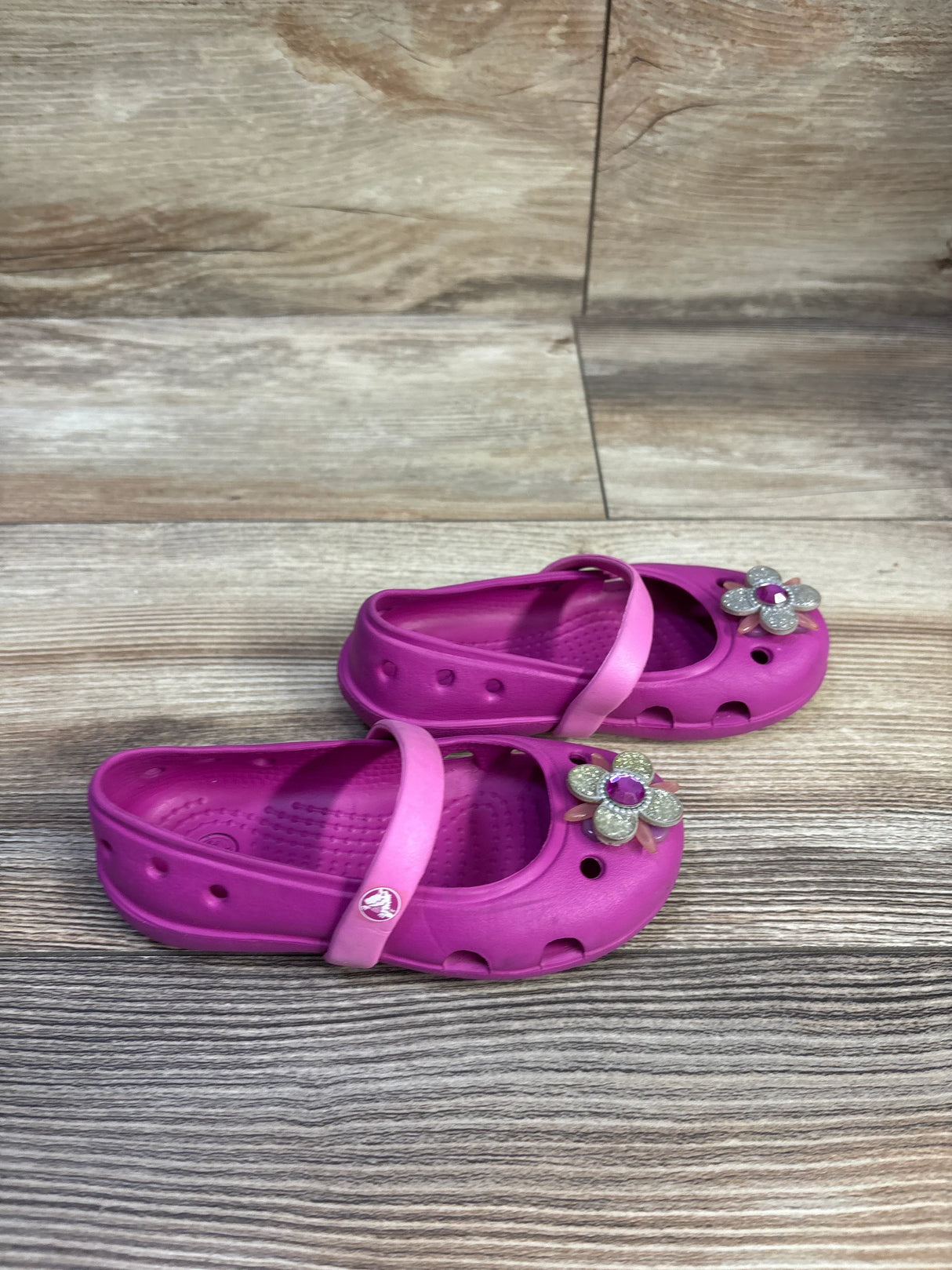 Three-quarter view of Crocs Keeley Petal Charm flats in pink showcasing flower charms and adjustable heel straps on wood floor background