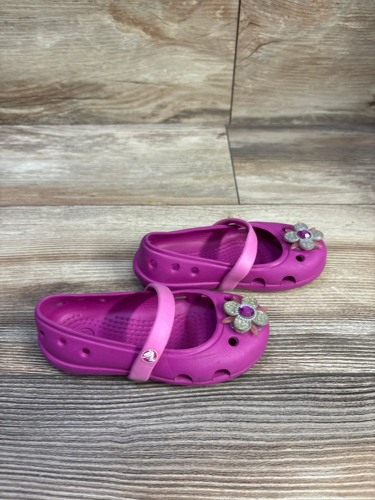 Three-quarter view of Crocs Keeley Petal Charm flats in pink showcasing flower charms and adjustable heel straps on wood floor background