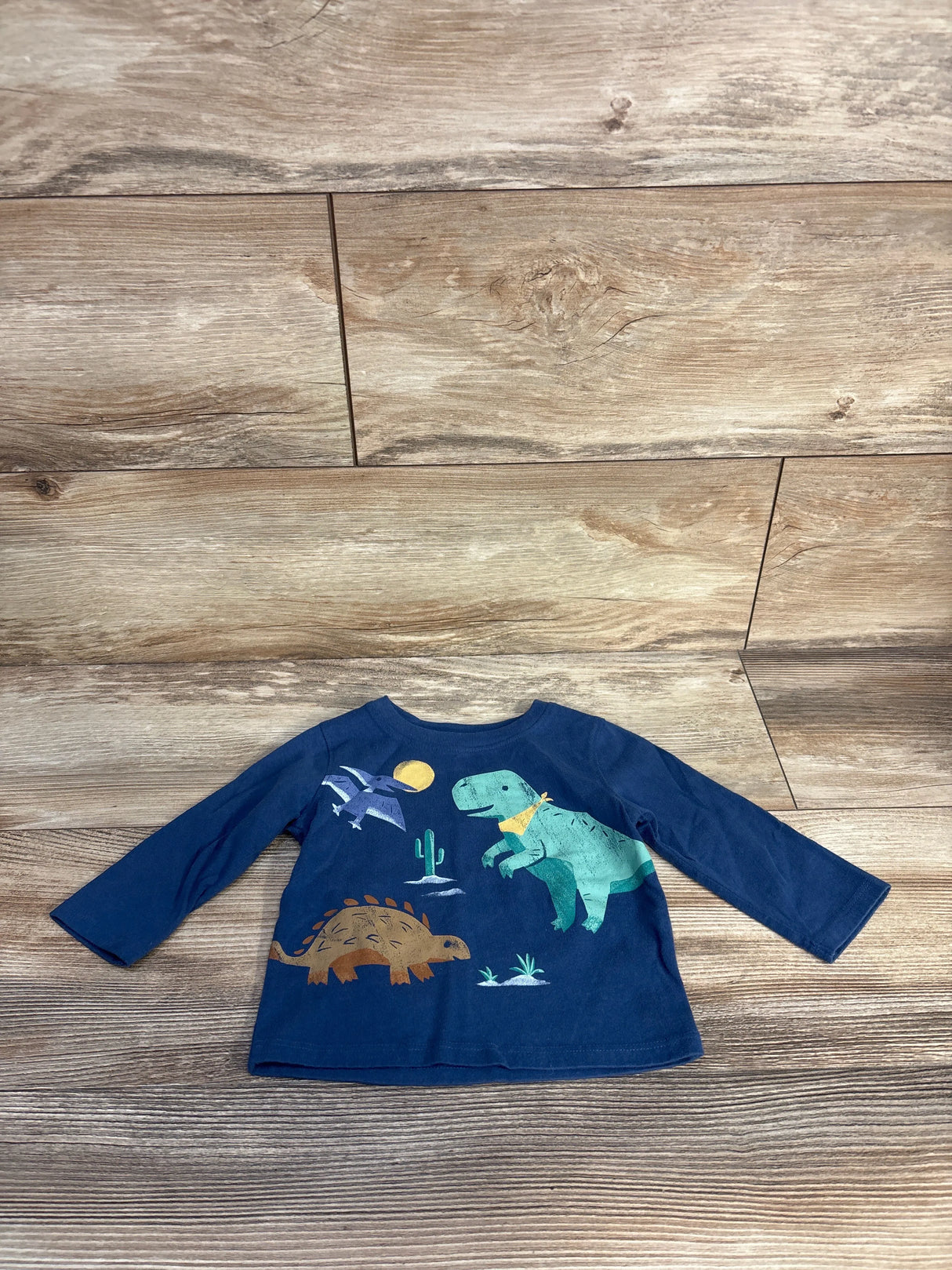 Flat lay of blue Jumping Beans long-sleeve baby shirt with dinosaur print (green T-rex, brown ankylosaur) on wood background