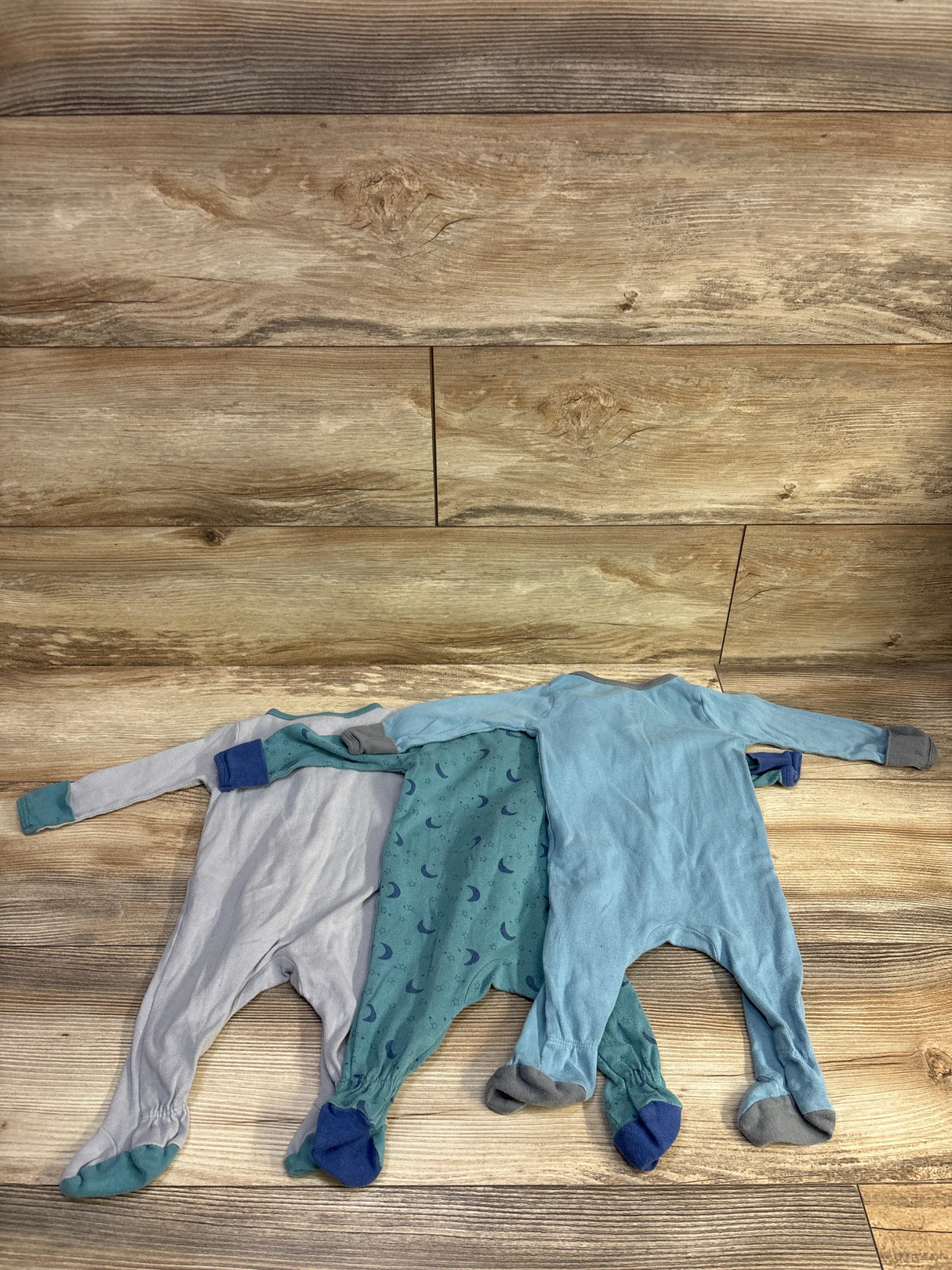 Flat-lay of Cloud Island 3-pack blue footed sleepers showing light blue, teal moon-print, and pale blue sleepers on wood backdrop